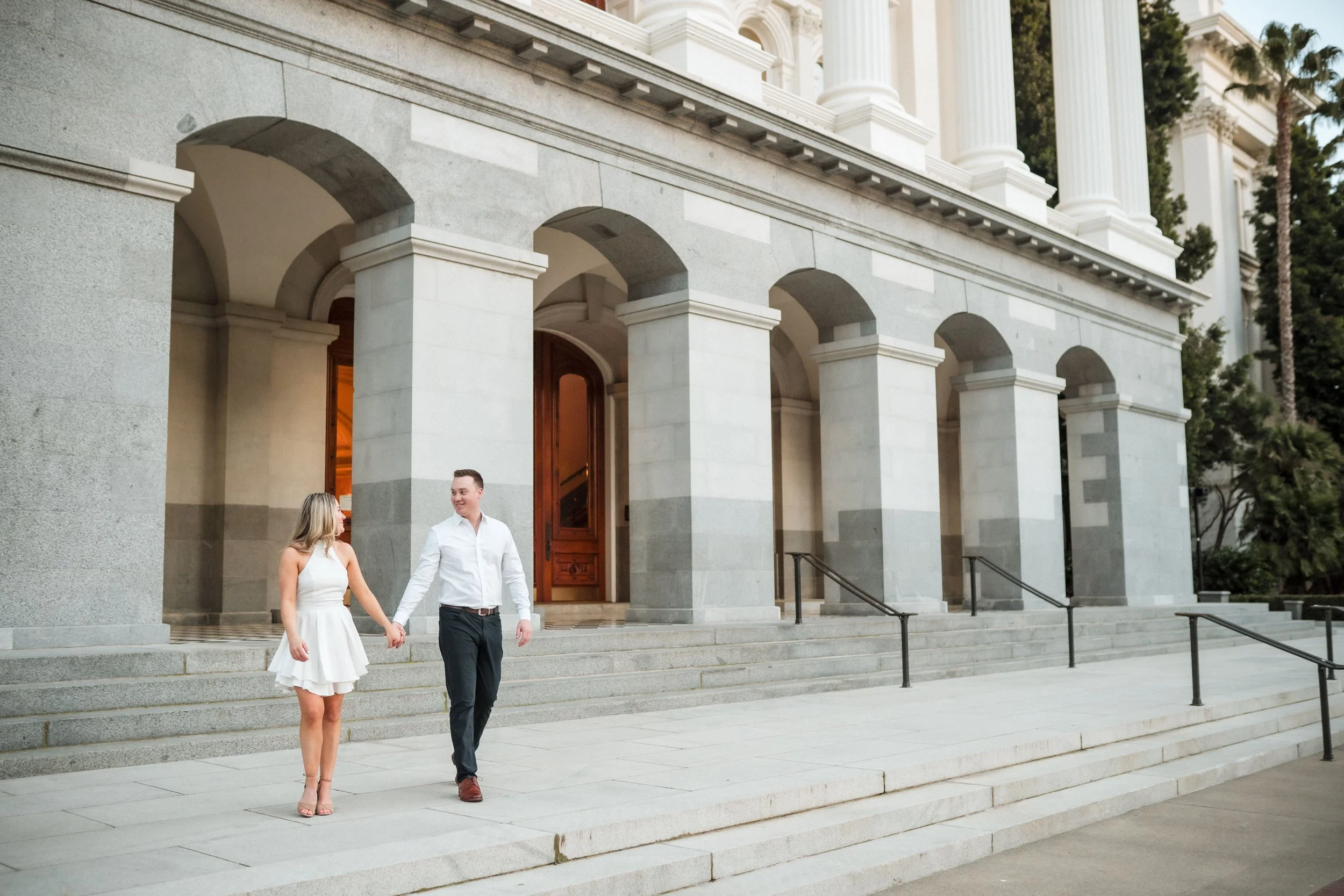 A couple holding hands walking down the steps of the Sacramento Capitol building, during their engagement session with Katherine Nicole Photography.