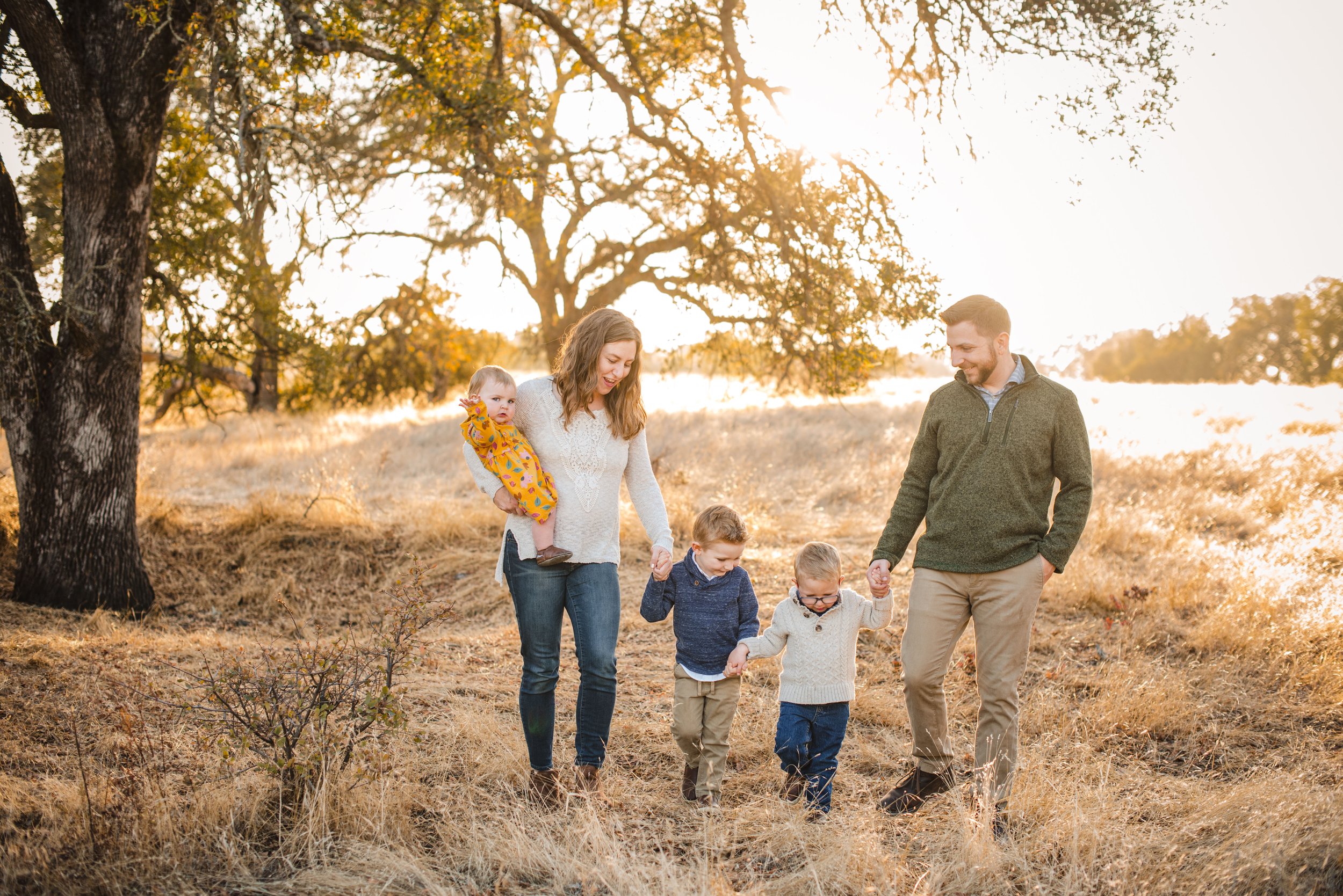 A happy family of five, two adults and three children, walking outdoors in a field with tall, golden grass, trees, and sunlight filtering through during family photos with Sacramento family photographer Katherine Nicole Photography. 
