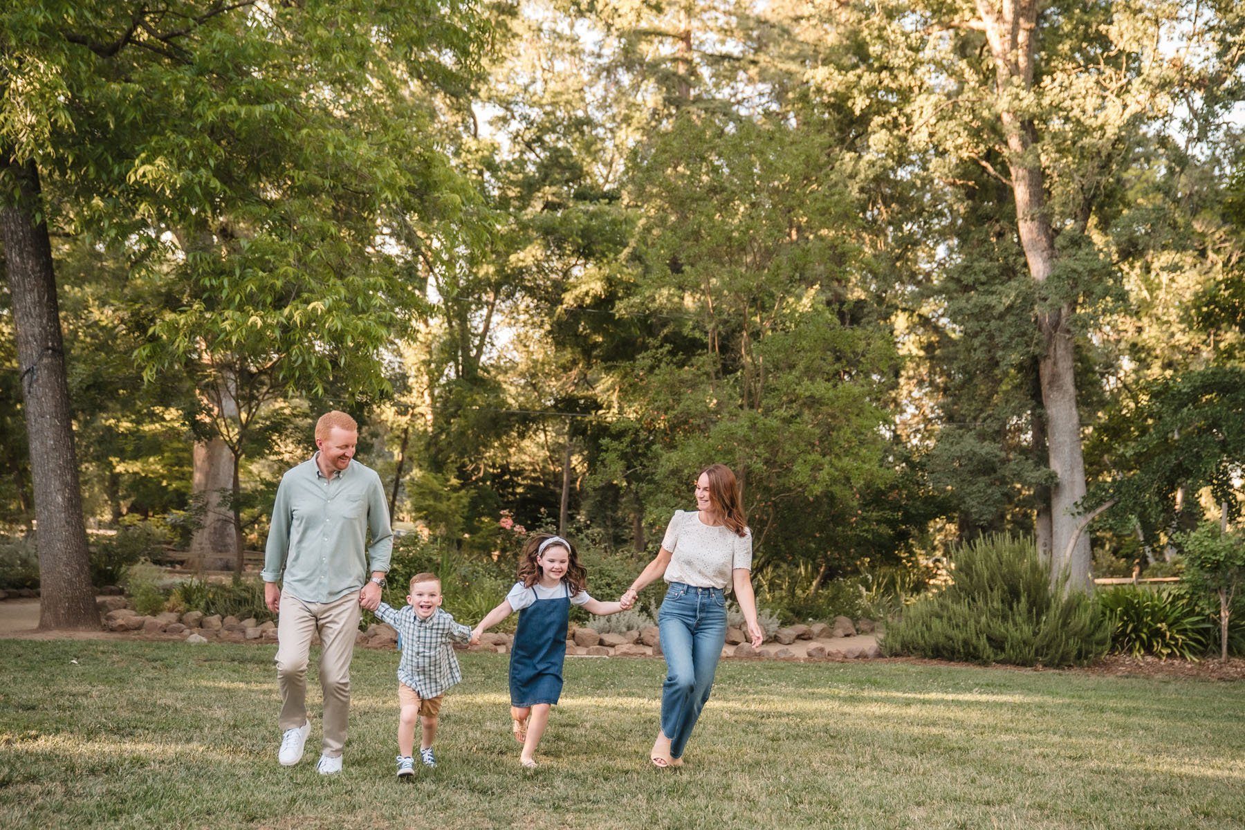 A happy family of five walking hand in hand in a park with tall trees and green grass during family photos with Sacramento family photographer, Katherine Nicole Photography.