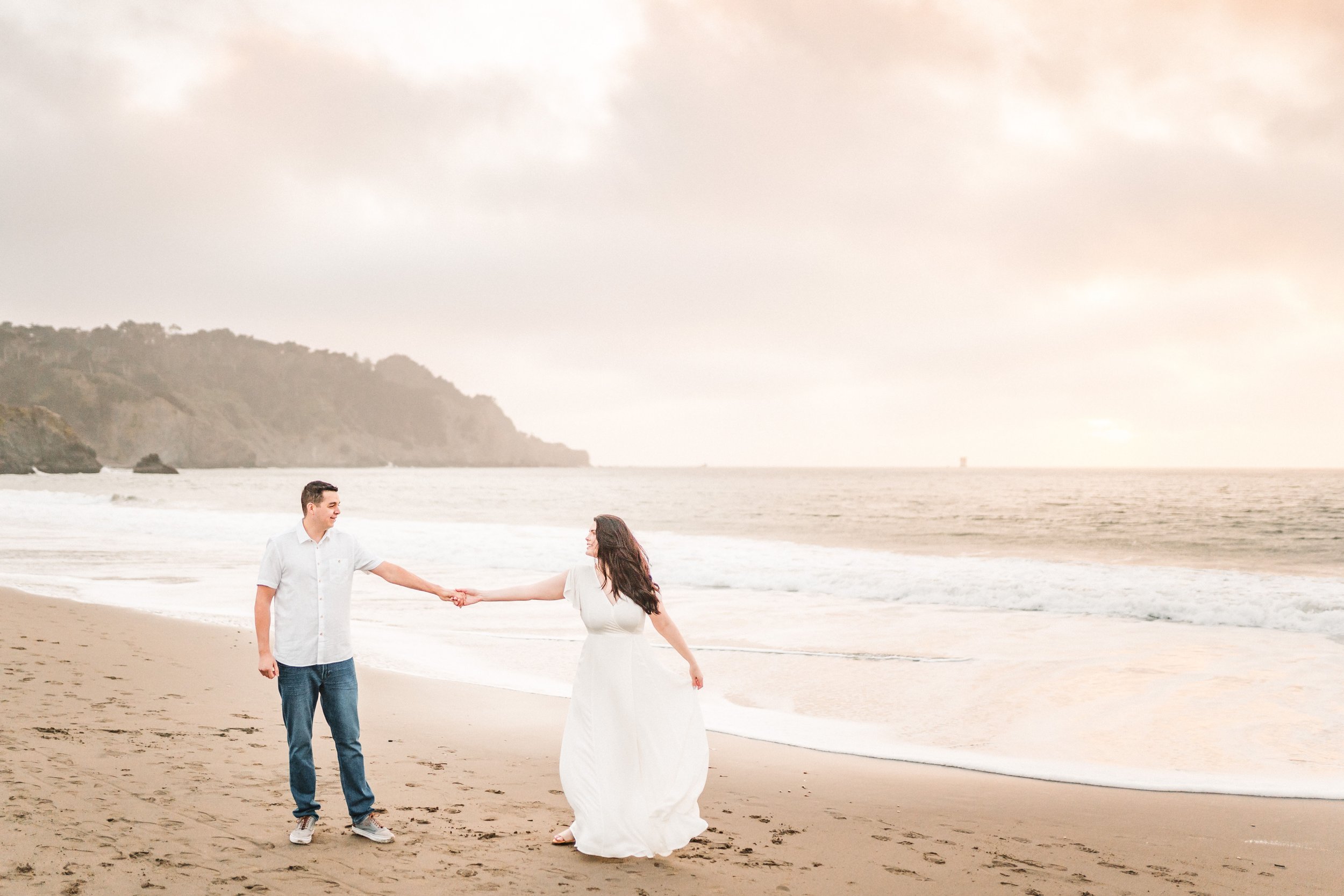 A man and woman holding hands on a beach, with waves and cliffs in the background, during their Bay Area engagement session. Photography by Katherine Nicole Photography.