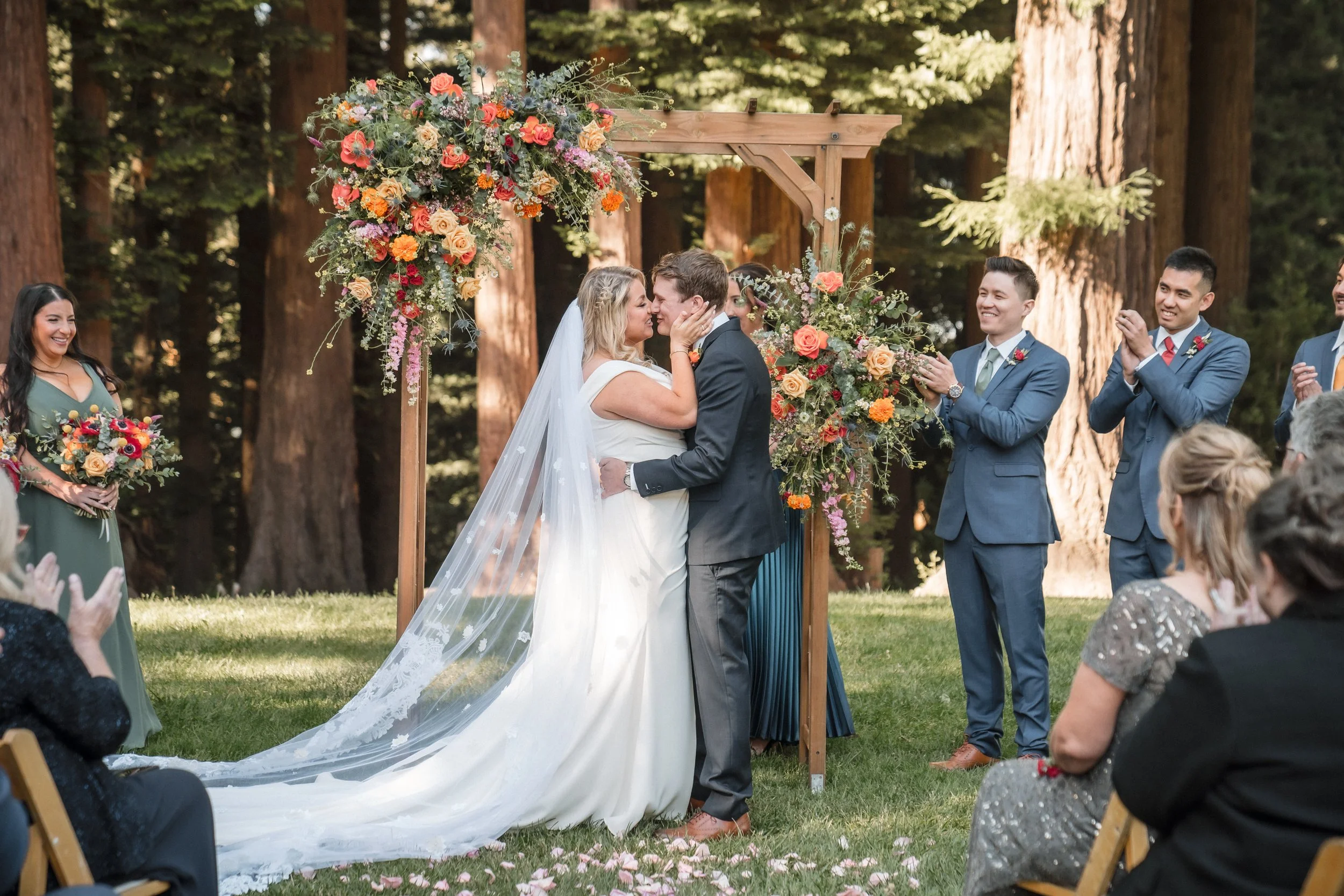 A couple kissing during their outdoor wedding ceremony at the Mountain Terrace, with a floral arch and seated guests in the background, photographed by Katherine Nicole Photography.