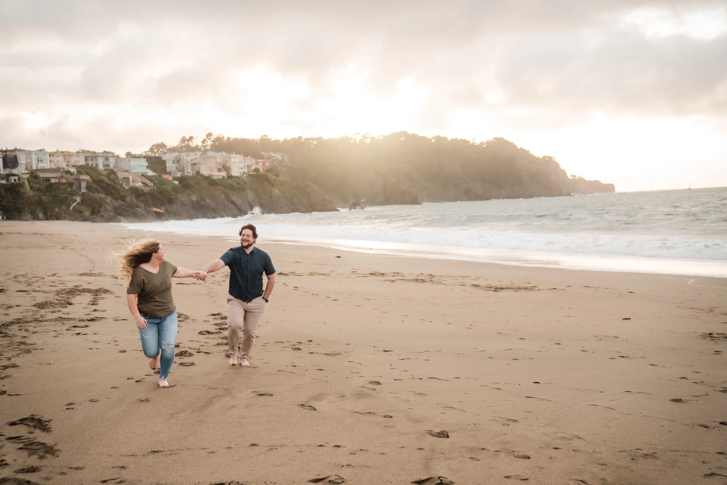 A man and woman holding hands and running barefoot on Baker Beach during their engagement session, with ocean waves and cliffs with houses in the background. Photography by Katherine Nicole Photography.