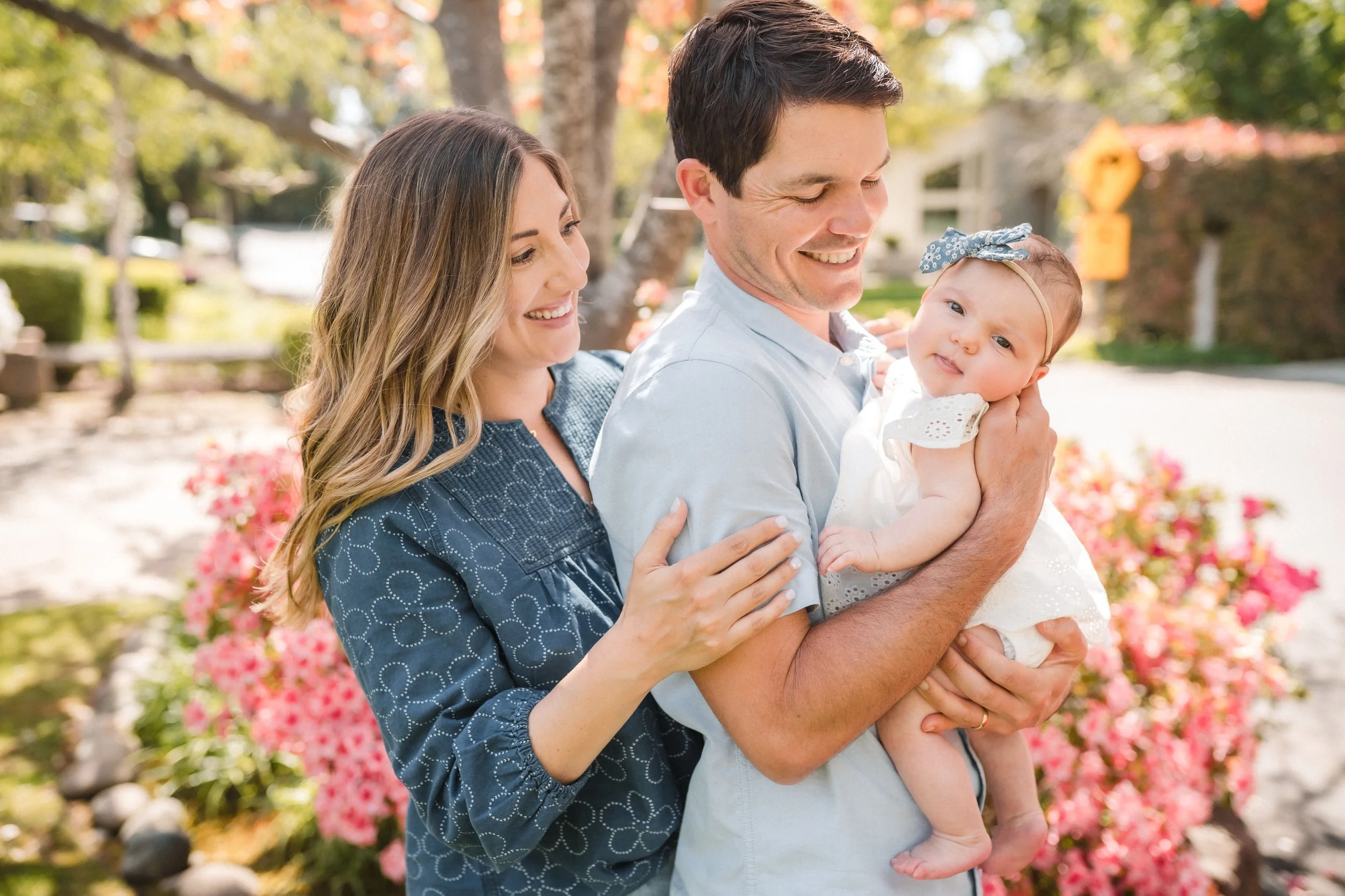 A family of three enjoying outdoor time on a sunny day, with a woman, man, and baby girl surrounded by flowers and trees, while taking family photos with Sacramento family photographer Katherine Nicole Photography. 