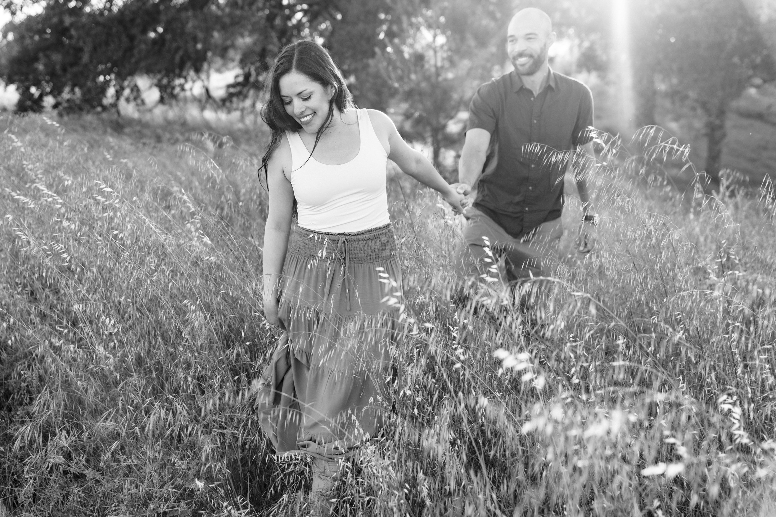 A black and white photo of a woman and a man holding hands while walking through a grassy field during their engagement session. The woman is looking down, while the man is smiling and looking at her. Photography by Katherine Nicole Photography.
