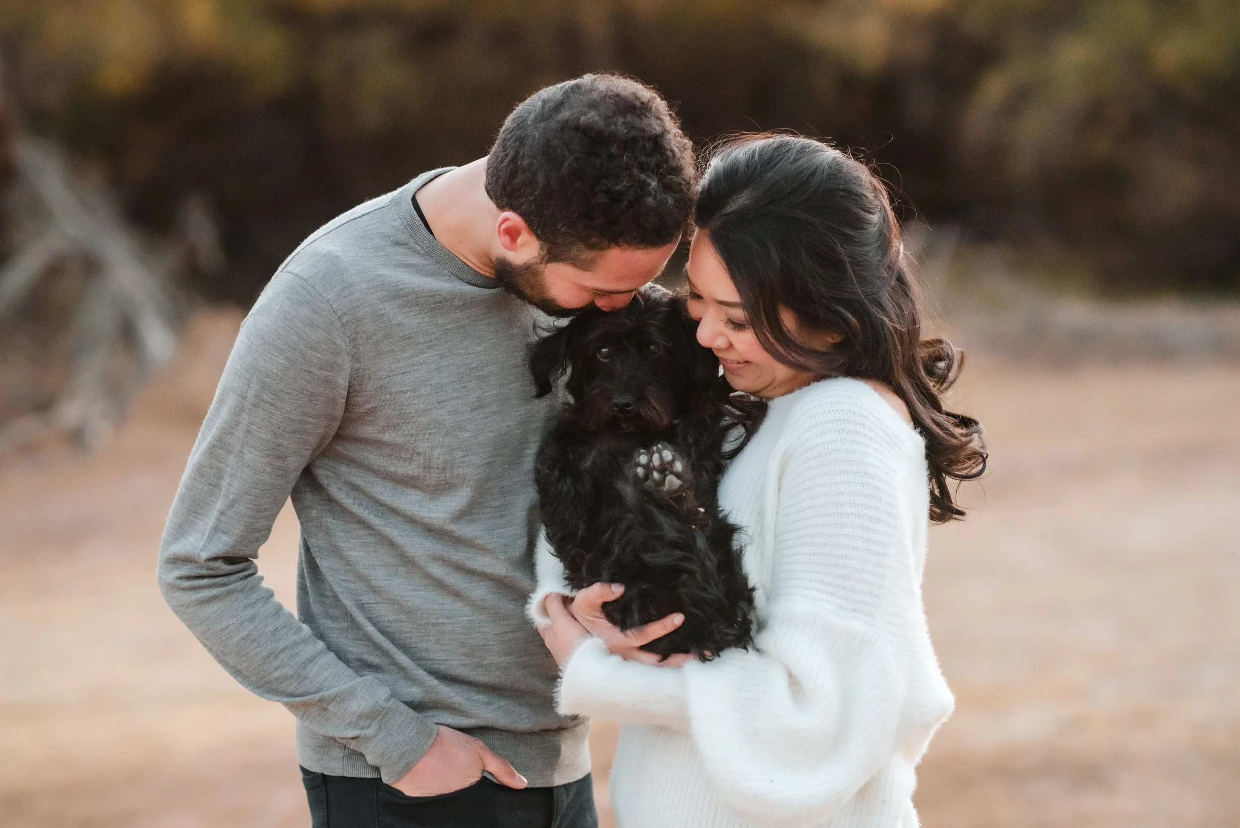 A couple lovingly holds their black puppy while leaning their foreheads together outdoors during their engagement session. Photography by Katherine Nicole Photography.