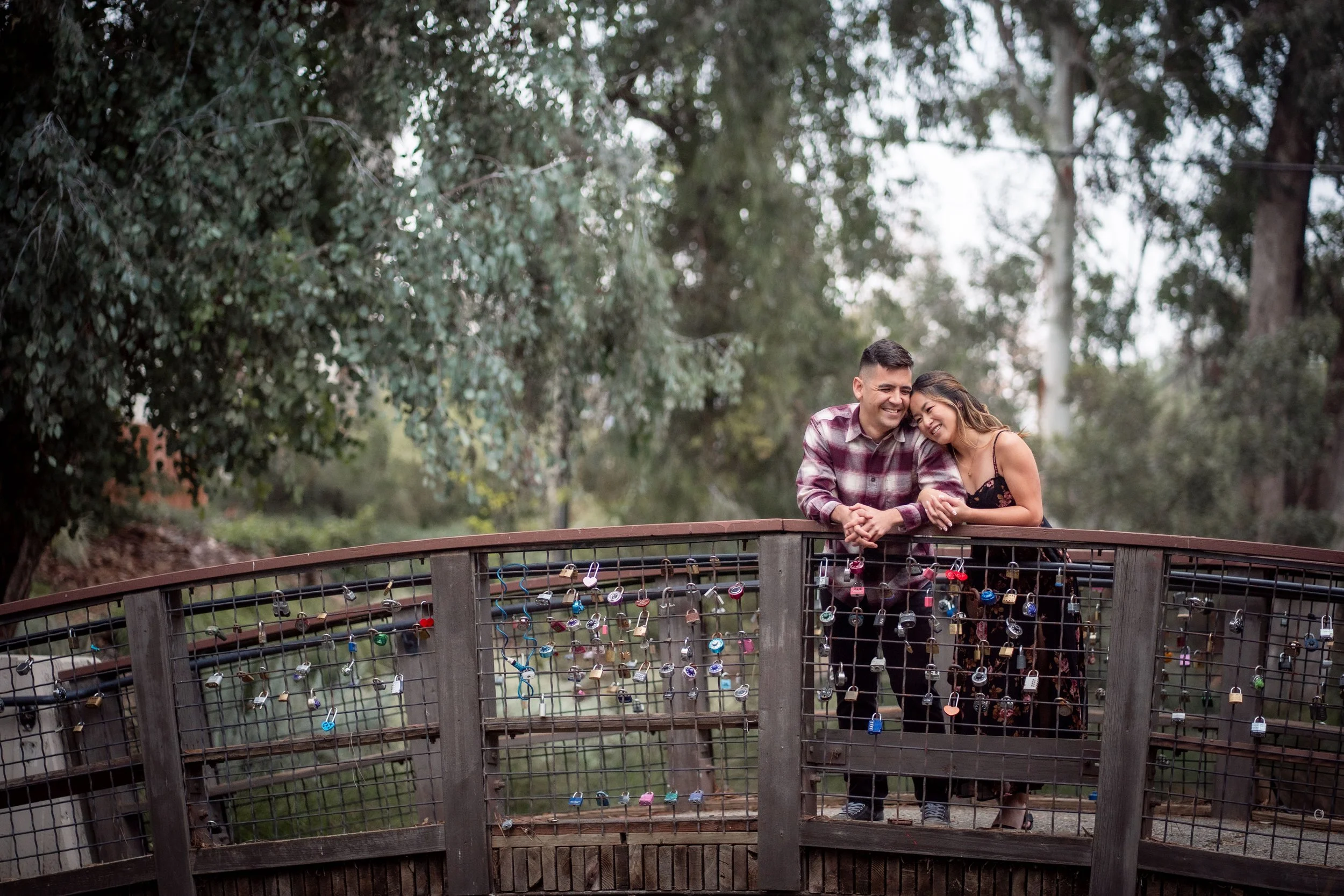 A smiling young couple leaning on a wooden bridge at the UC David Arboretum, railing adorned with colorful padlocks, surrounded by tall trees at their engagement session. Photography by Katherine Nicole Photography.