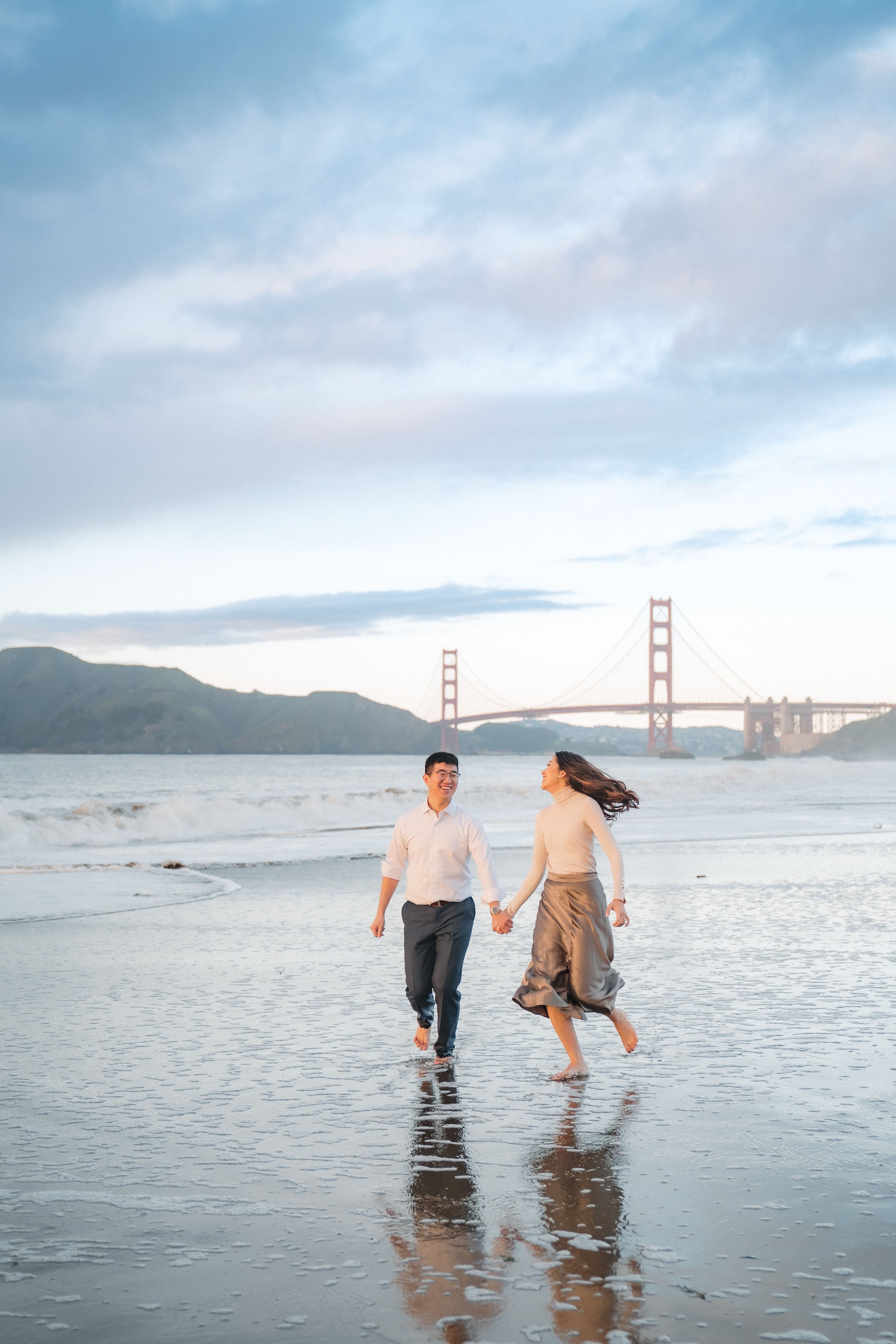 A couple walking hand in hand on a beach near the water, with the Golden Gate Bridge and hills in the background during sunset while taking their engagement photos with Katherine Nicole Photography.