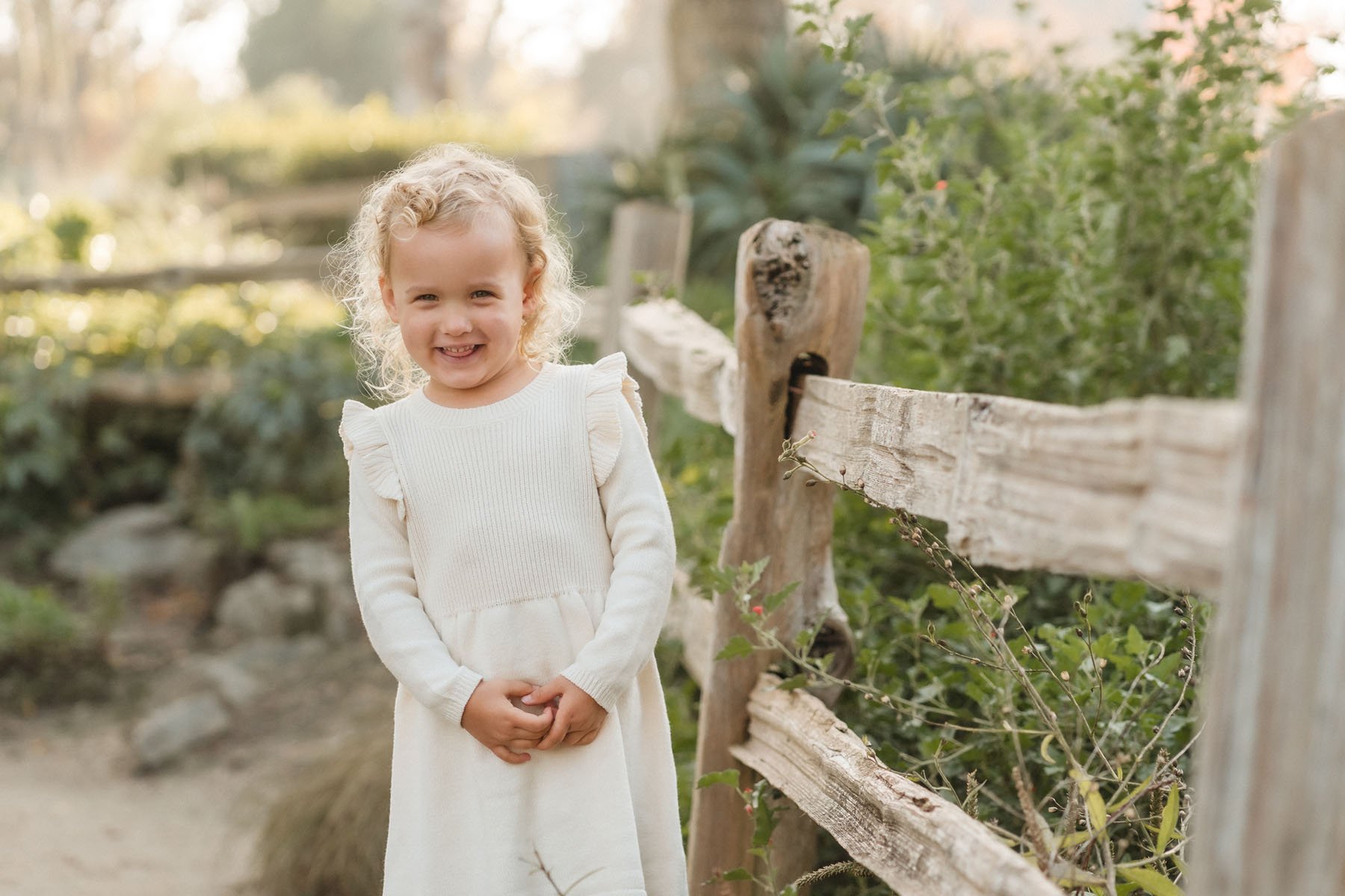 A young girl with blonde curly hair, smiling, standing outdoors next to a rustic wooden fence with greenery in the background. Photography by Katherine Nicole Photography.