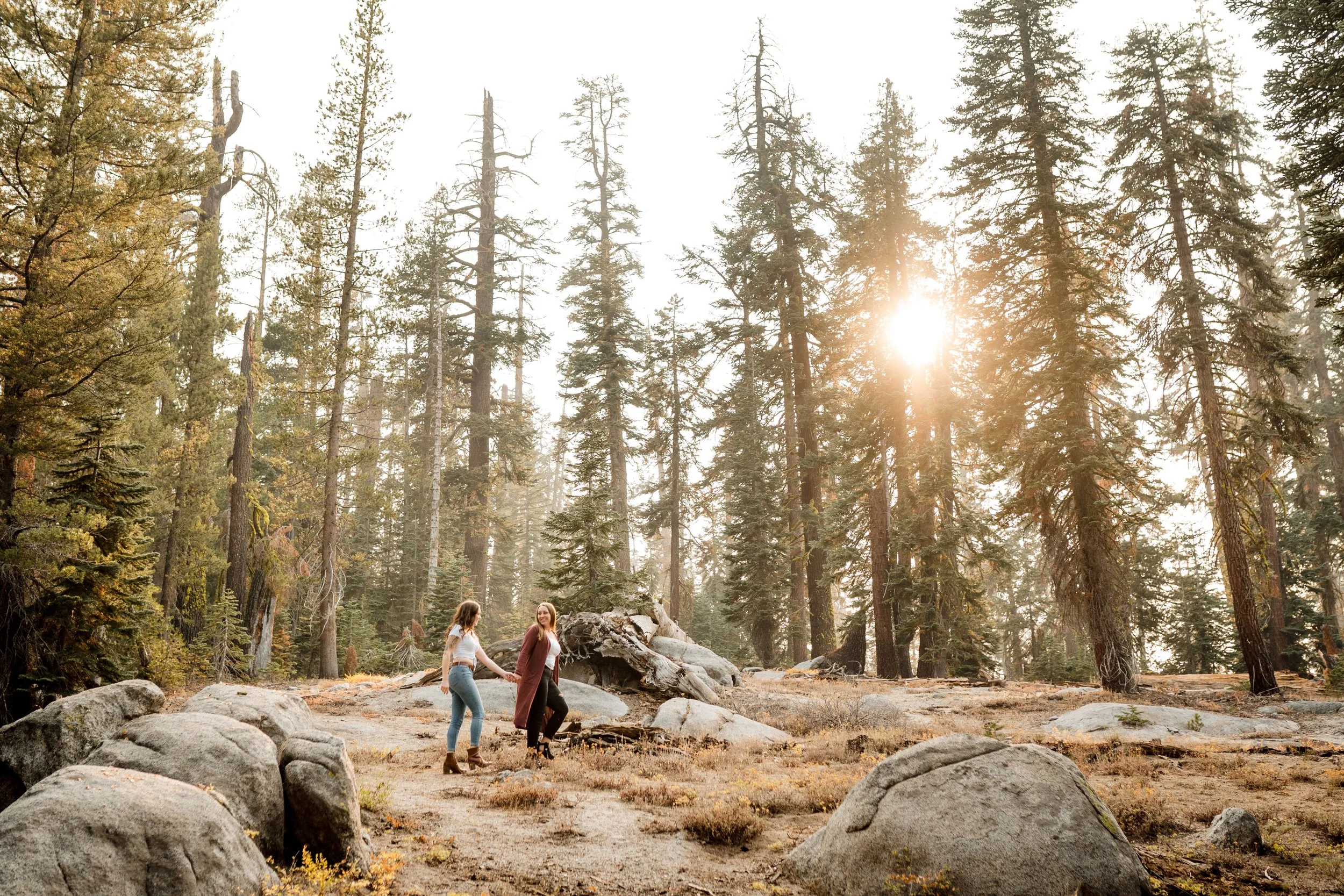 Two women are walking hand in hand through Yosemite near Taft Point, with tall pine trees and large rocks, in a natural outdoor setting during their Yosemite engagement session. Photography by Katherine Nicole Photography.