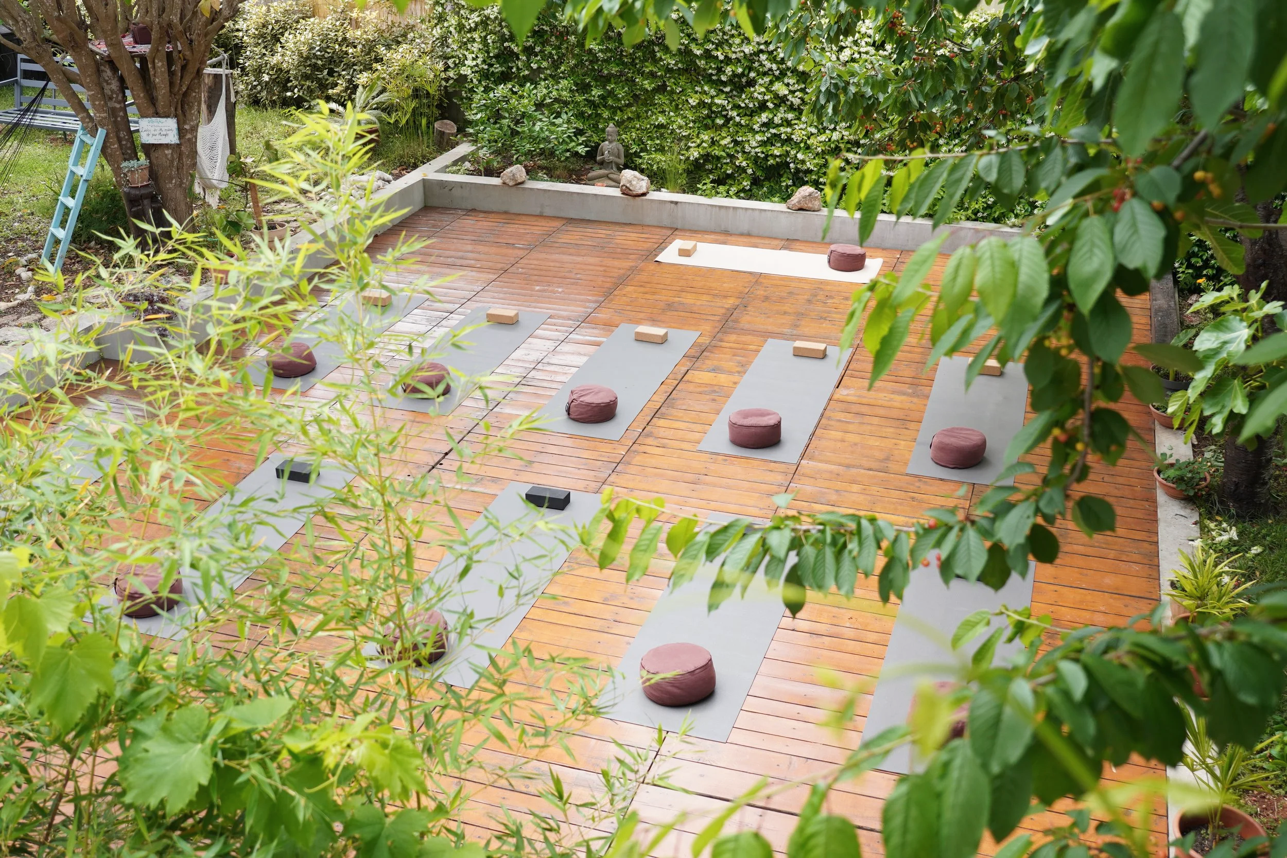 A tranquil outdoor yoga space on a wooden deck surrounded by lush green plants and trees. Multiple yoga mats with cushions are arranged on the deck, with some mats on a white mat or a grey mat. A small statue of a seated Buddha is visible in the background, along with potted plants and rocks, creating a peaceful garden setting.