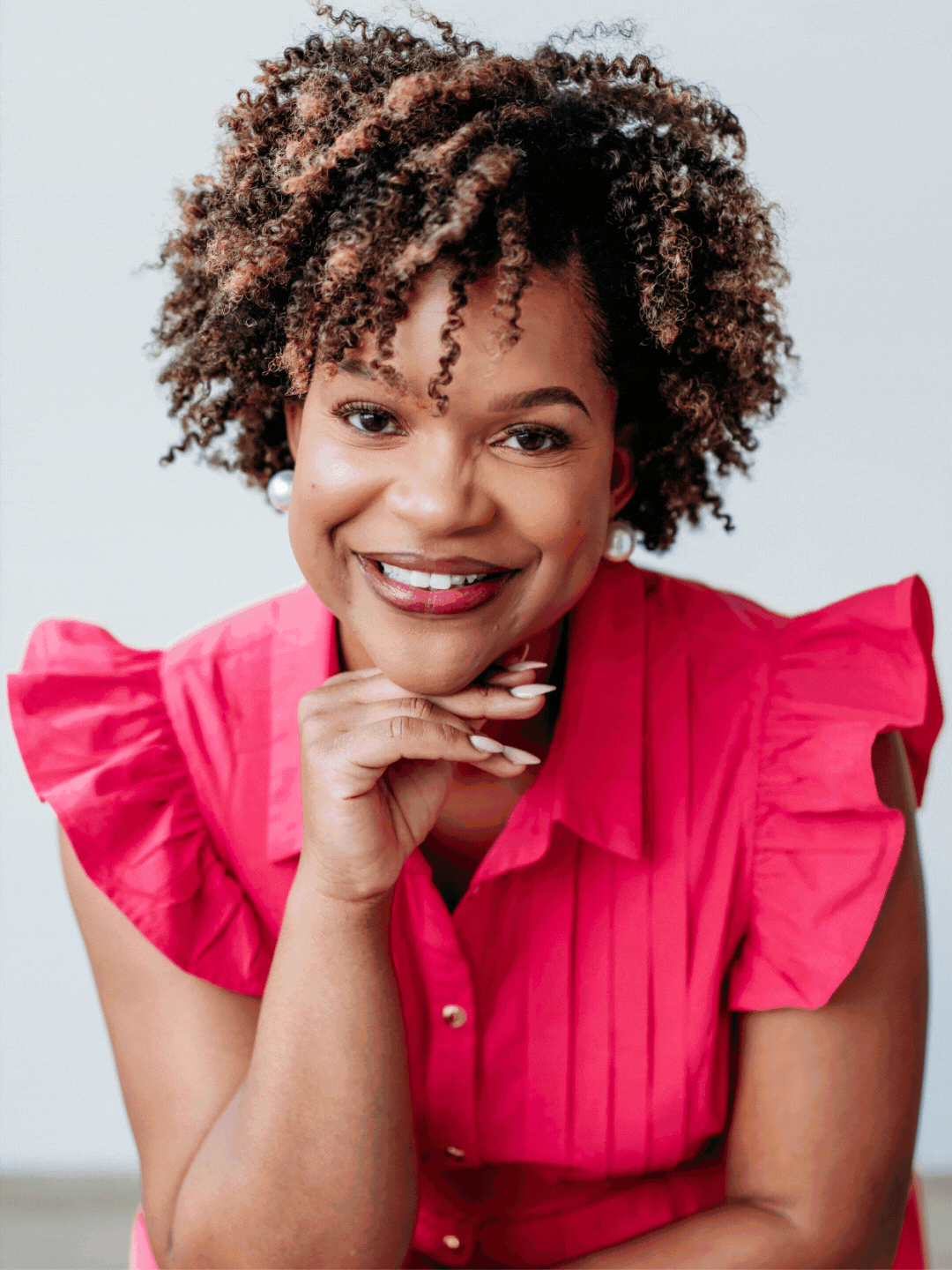 A woman with curly hair wearing a bright pink blouse with ruffled sleeves, smiling and resting her chin on her hand.