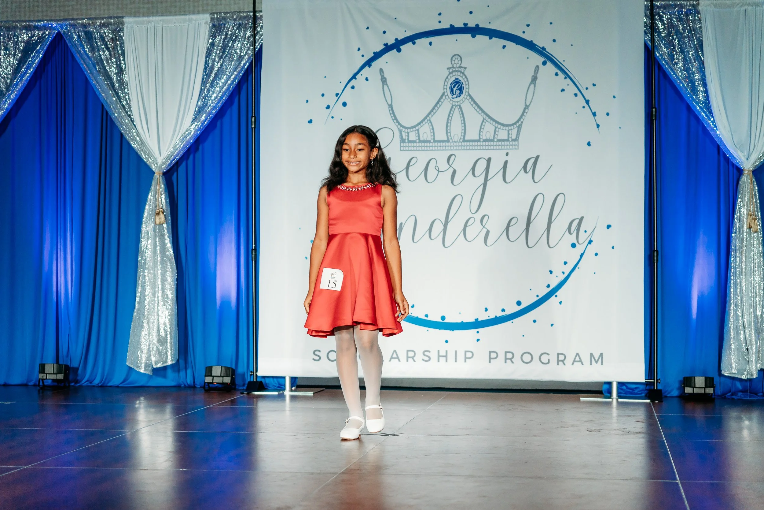A young girl in a red dress walking on stage during a scholarship program. She has a number 15 badge on her dress and is smiling. The backdrop has a crown illustration and the words 'Georgia Cinderella Scholarship Program' with blue and silver curtai