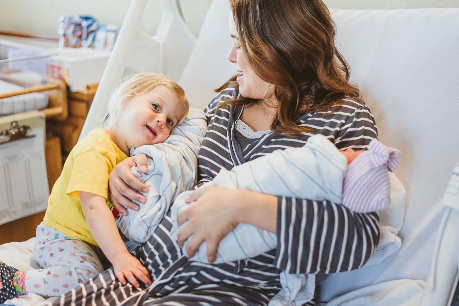 mom and toddler hugging newborn twin girls in hospital bed