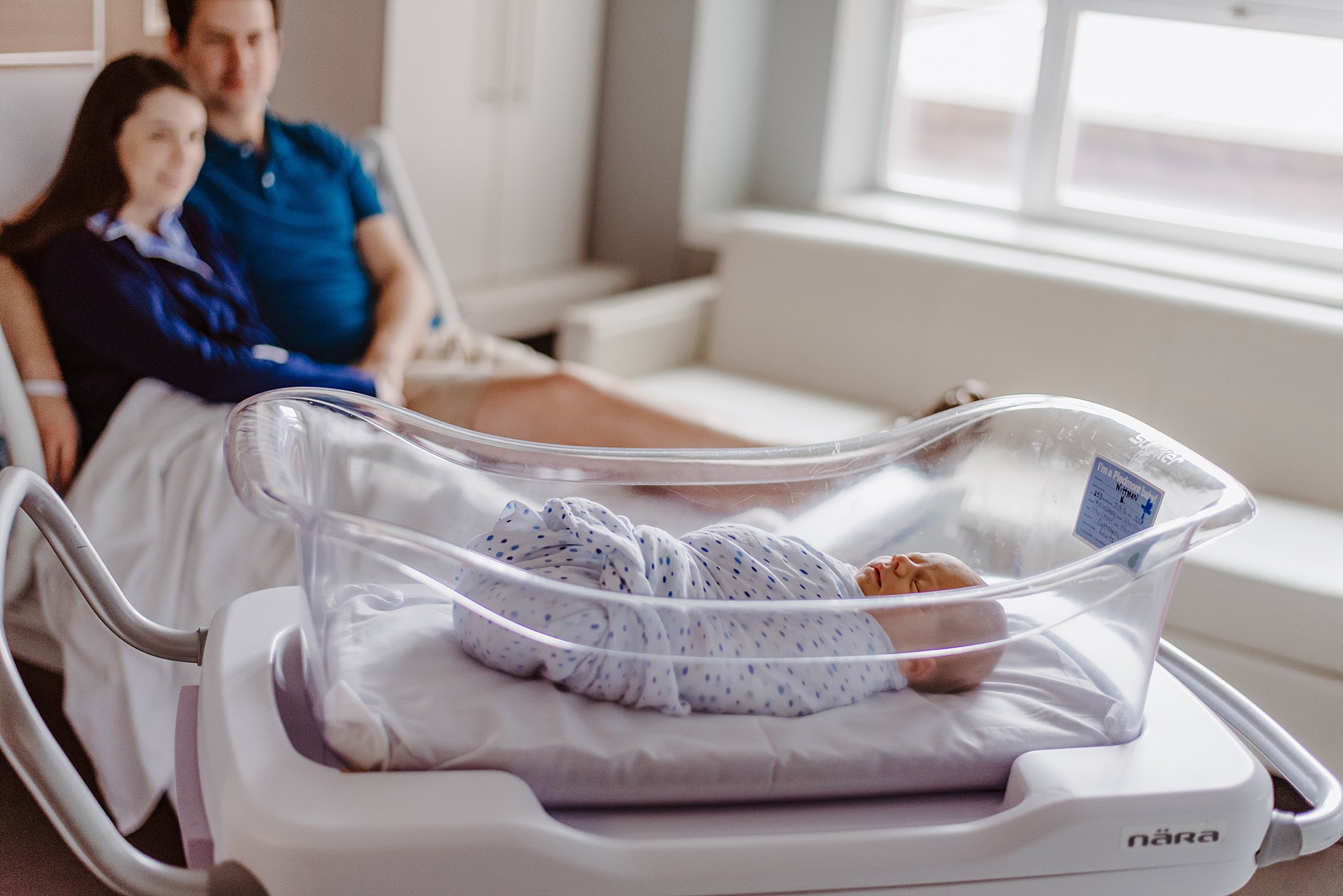 newborn baby boy in hospital bassinet with parents in background