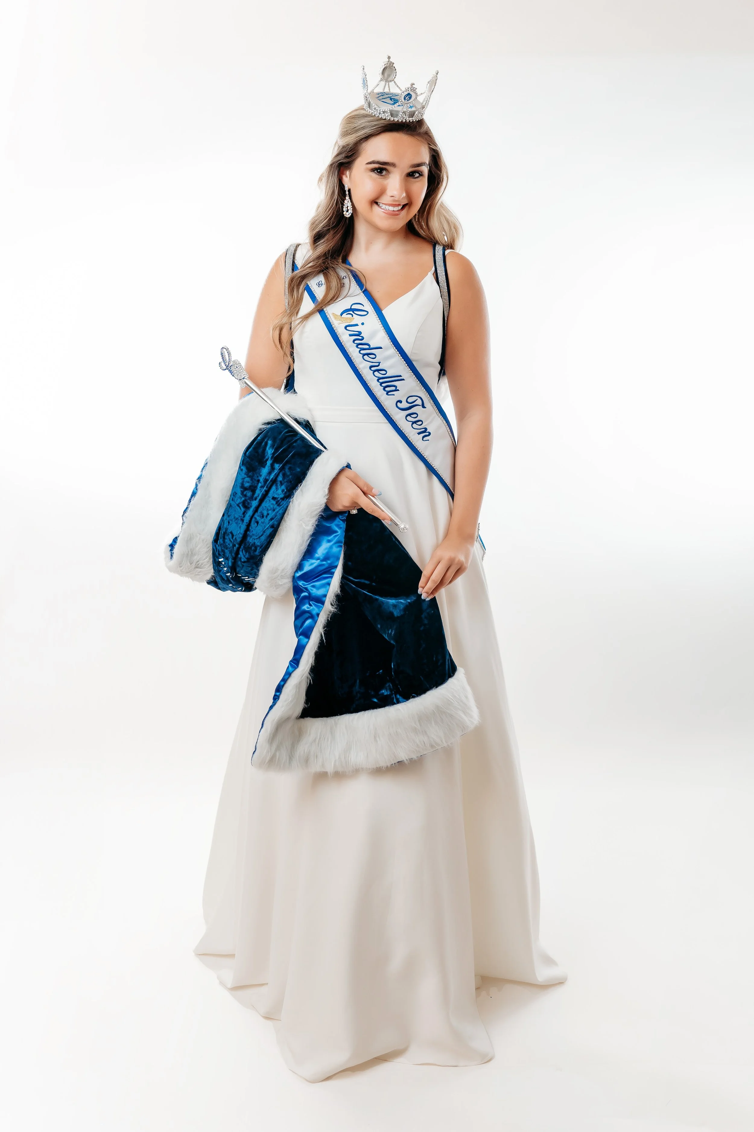 Young woman in a white dress wears a sash that says 'Gindarella Teen,' a crown, and holds a blue velvet cape with white fur trim.
