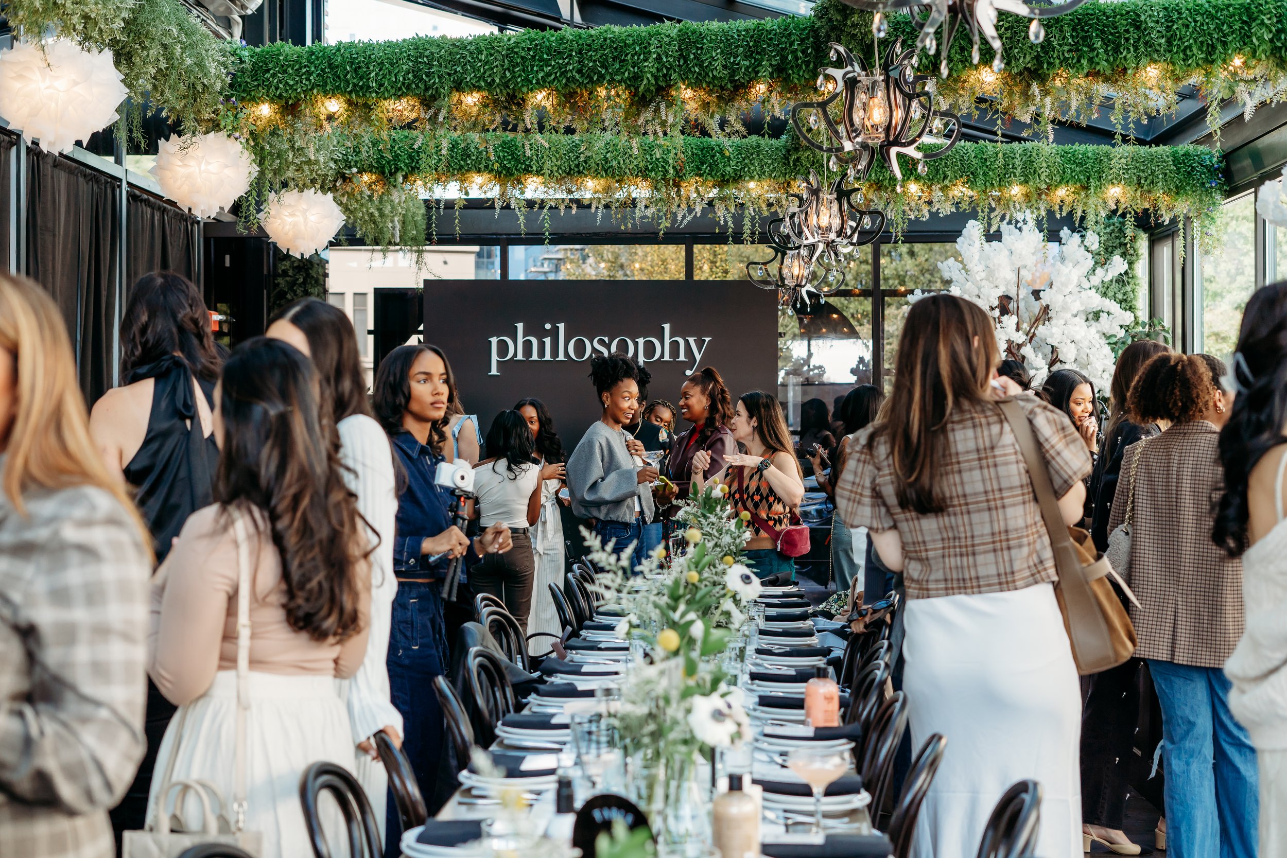 A group of women standing near a long dining table with floral decorations, chatting and smiling.