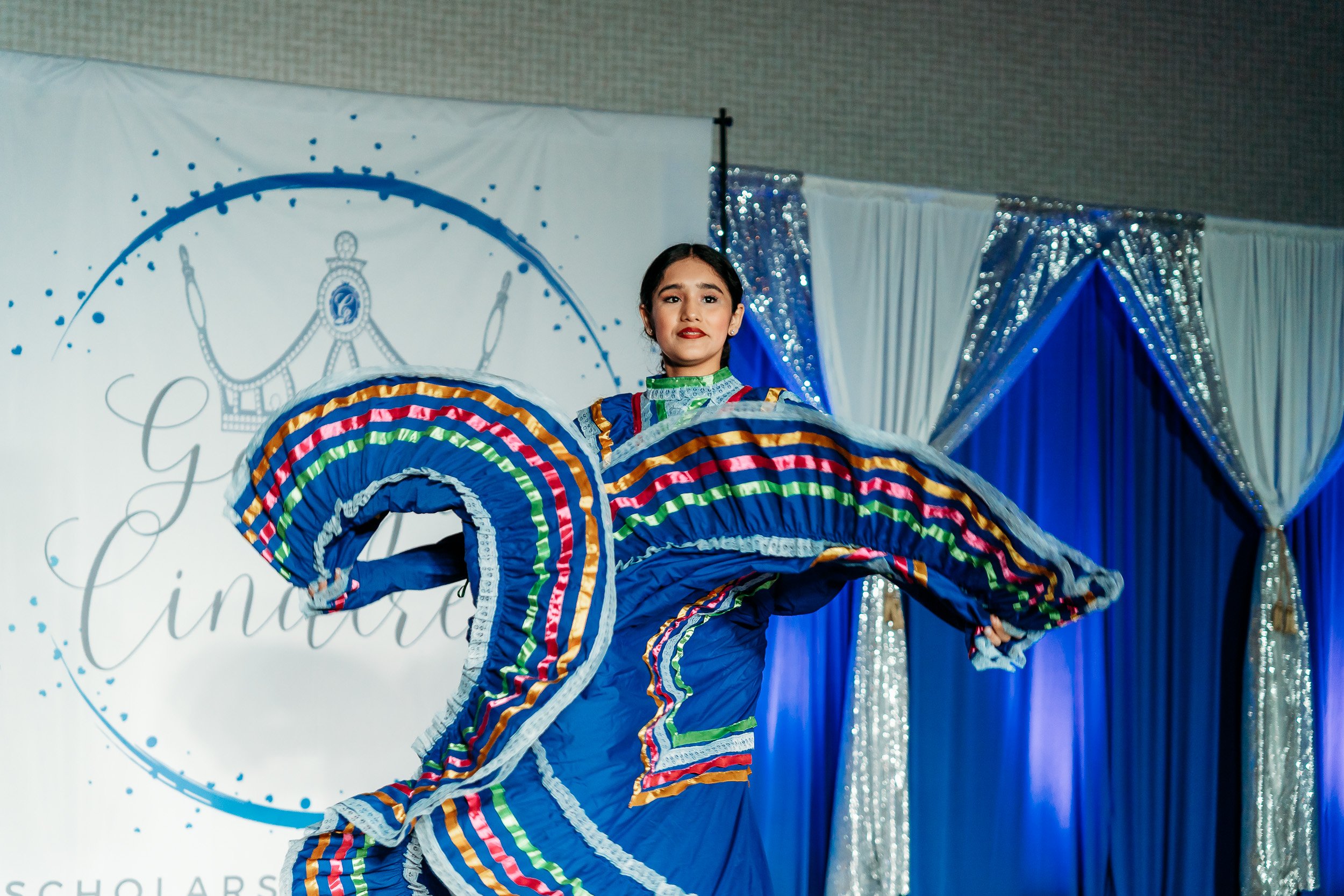 Young girl performing a traditional dance in a colorful dress with blue, green, red, and yellow stripes, on stage with a backdrop and blue curtains.