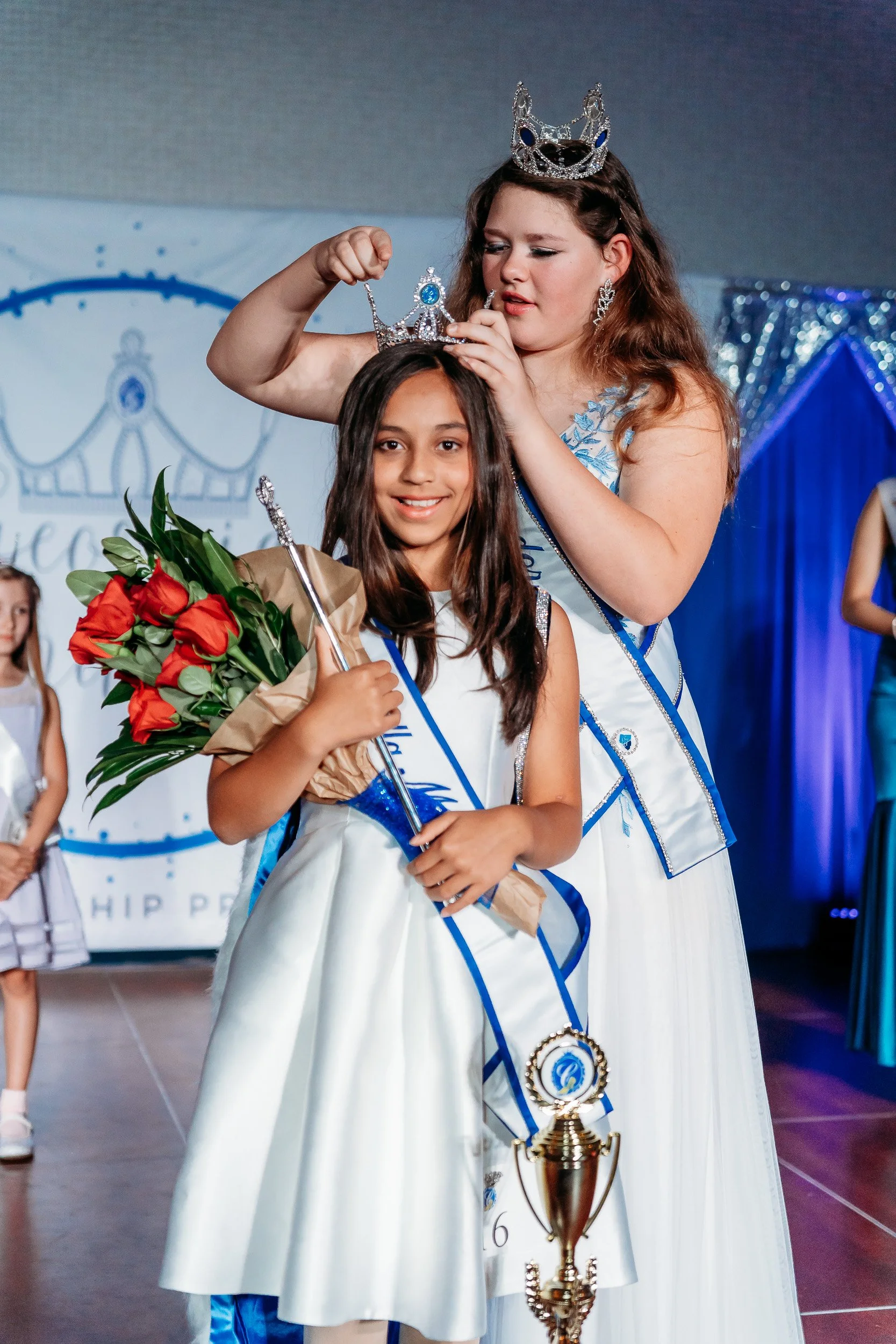 A young girl receiving a crown and sash at a beauty pageant, holding a bouquet of red roses and a small trophy, on stage with other contestants in the background.