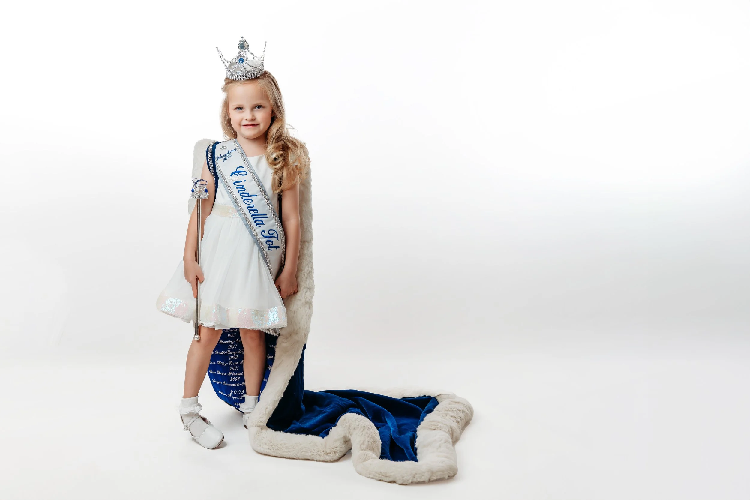 Young girl dressed as a beauty queen, wearing a crown, sash, and dress, standing next to a blue velvet royal cape with white fur trim, in front of a white background.