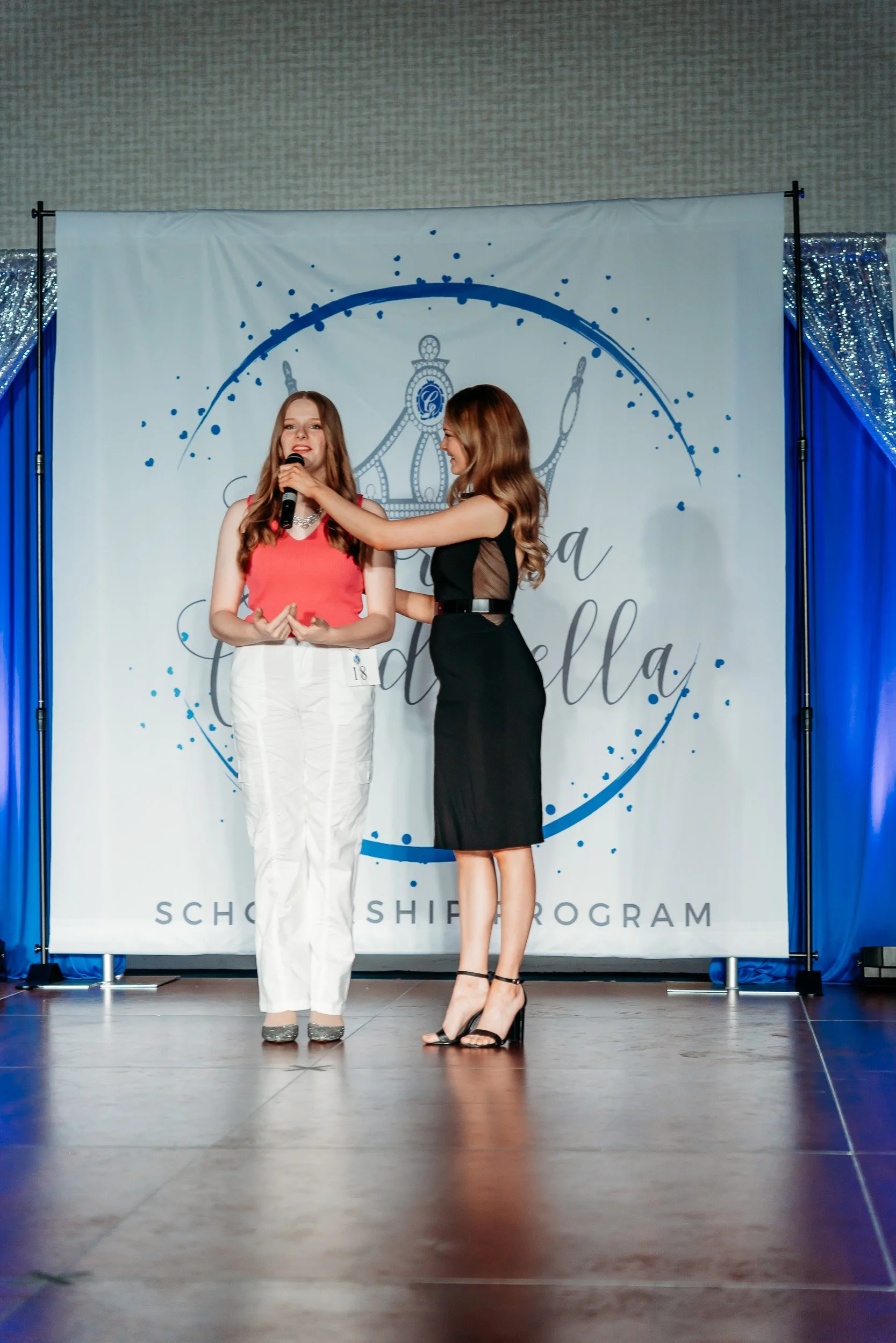 Two women on stage at a scholarship program event, one wearing a red top and white pants, being interviewed by the other in a black dress, with a backdrop that says "La Bella" and a crown illustration behind them.