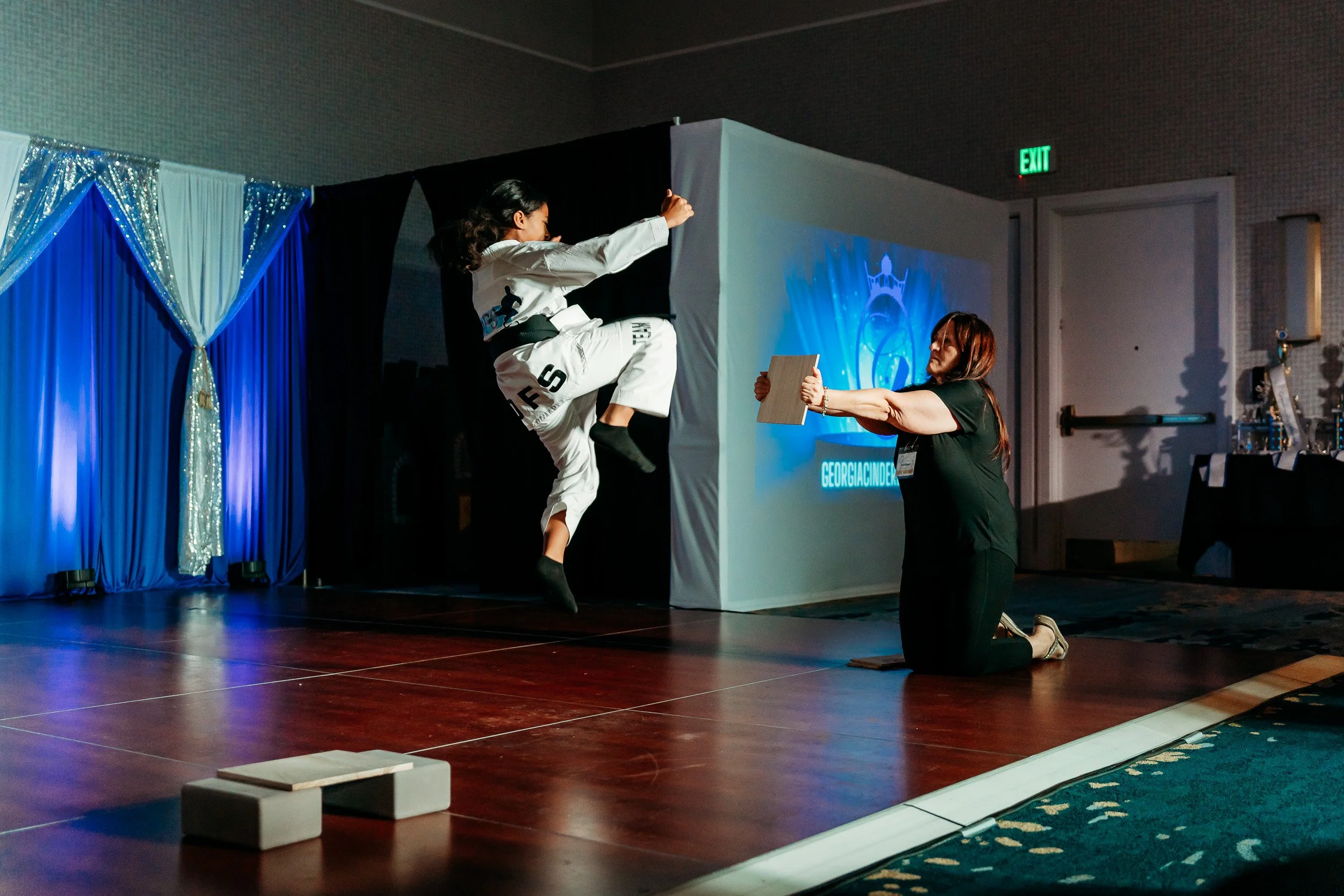 A woman is performing a martial arts move on stage, while another woman kneels on one knee, holding a wooden board. TheStage has blue and silver drapery with a projected logo in the background.