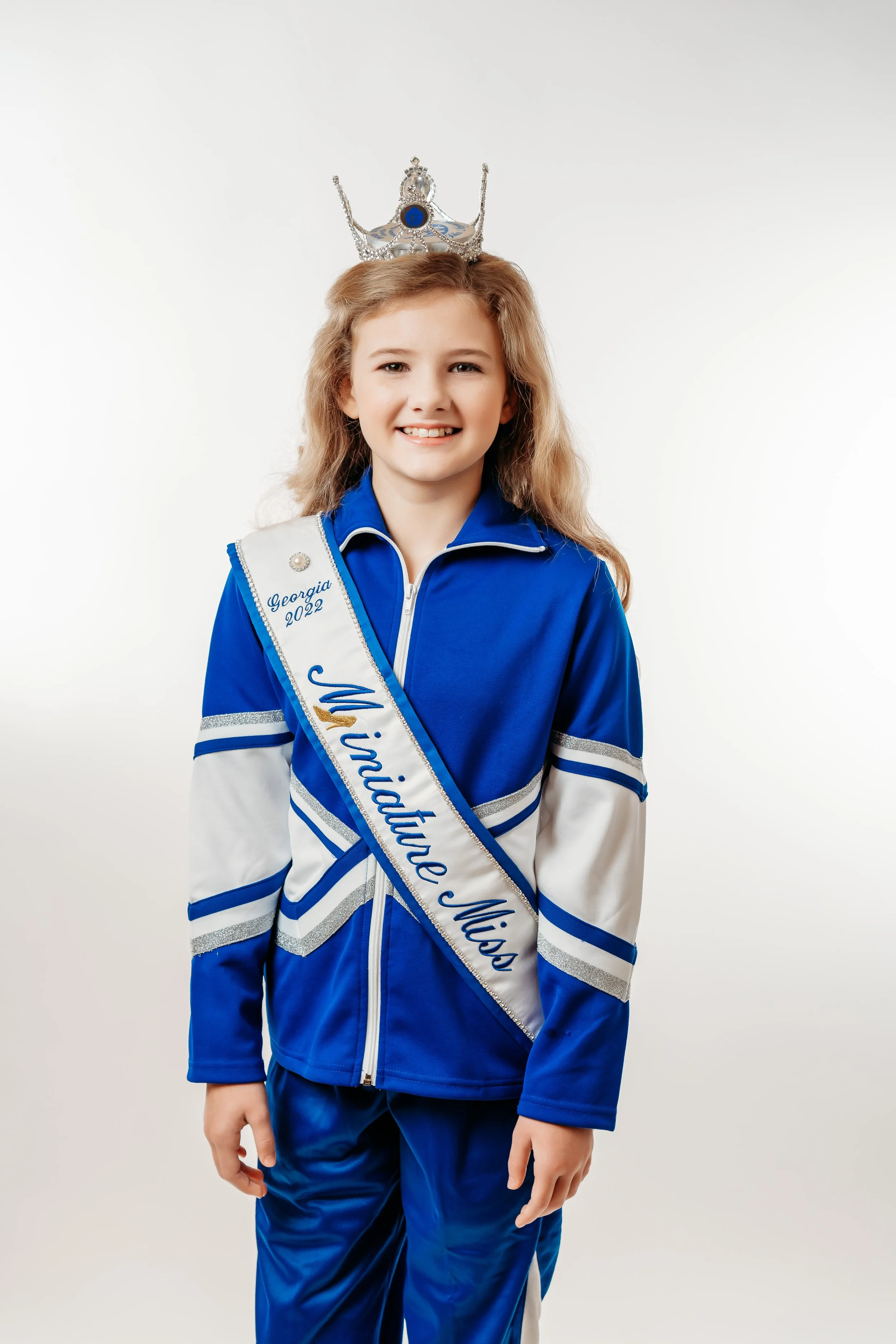 Young girl wearing a blue and white cheerleading outfit with a sash that reads 'Miss Midlothian' and a tiara on her head.