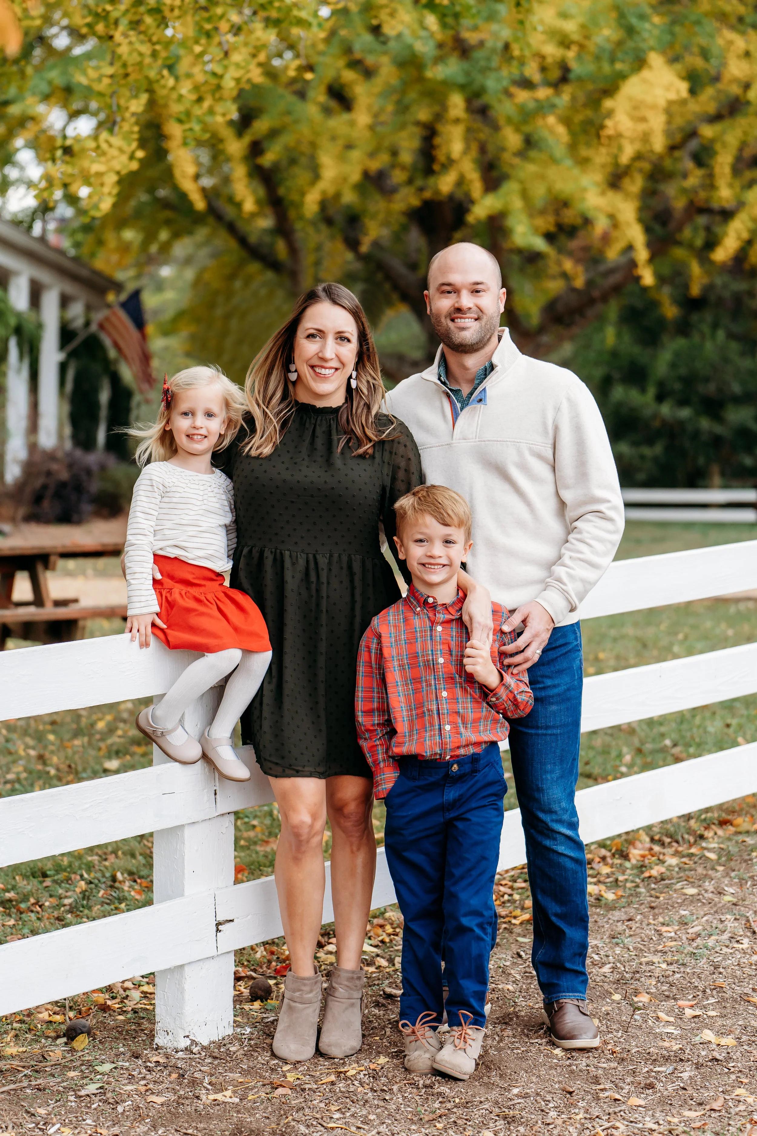 A family of four standing outdoors in front of a white fence with fall trees in the background. The mother, father, young girl, and young boy are smiling at the camera, dressed in casual clothes suitable for autumn.