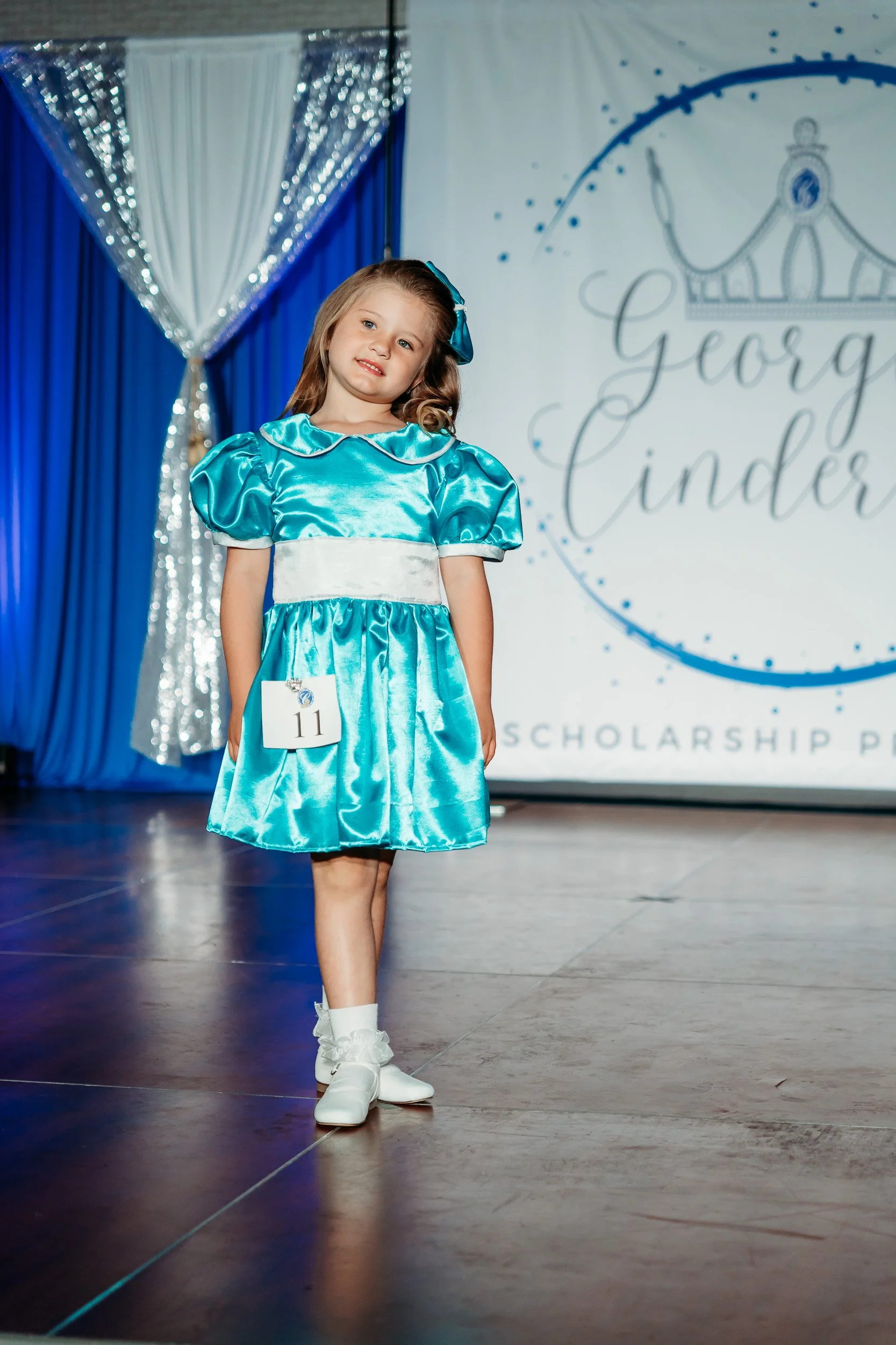 Young girl participating in a scholarship pageant, wearing a blue satin dress with puffed sleeves and white sash, standing on stage with a backdrop displaying her name, Georgiinder.