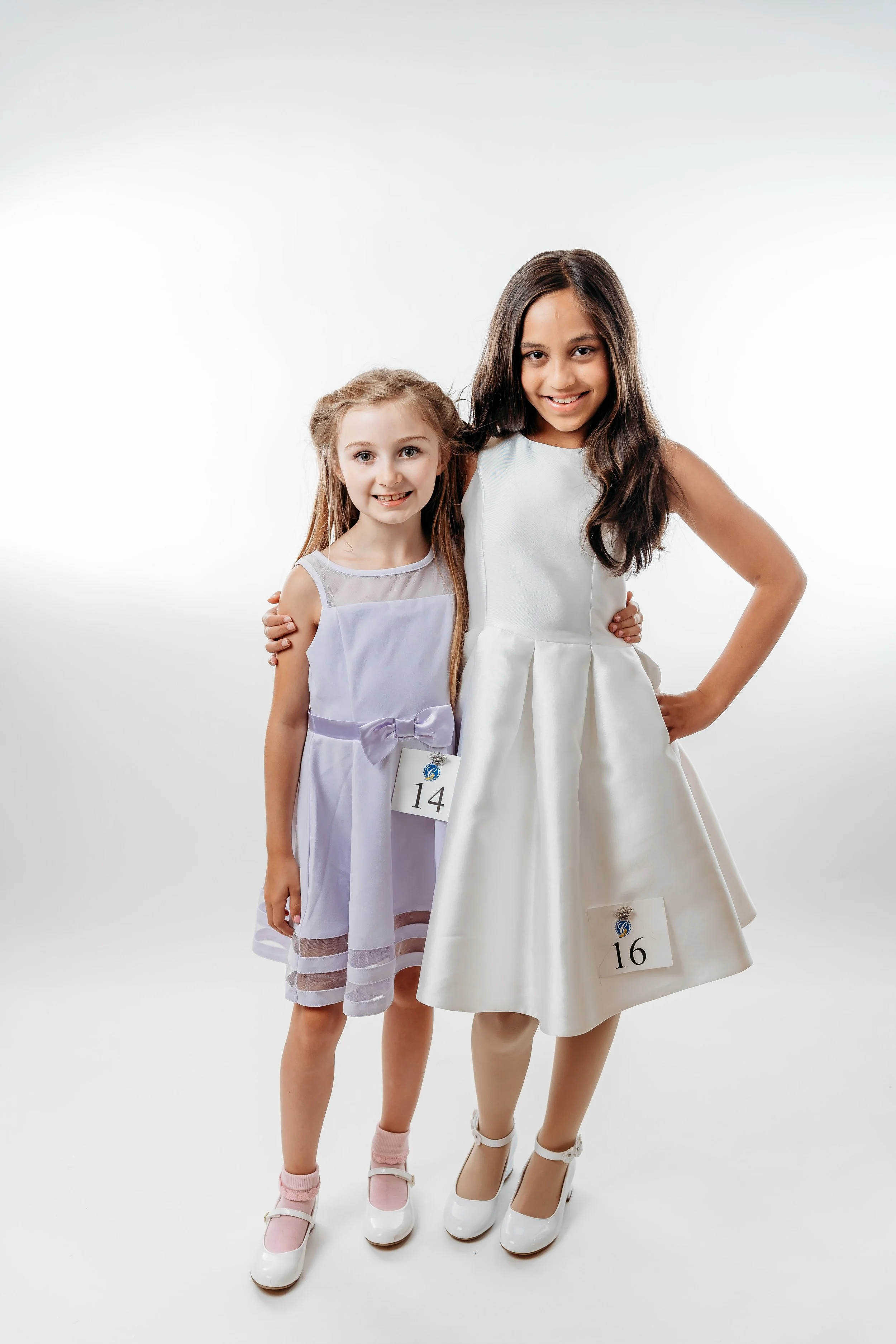 Two young girls dressed in formal attire, standing together against a white background at a pageant or event, each with a number badge pinned to their dresses.