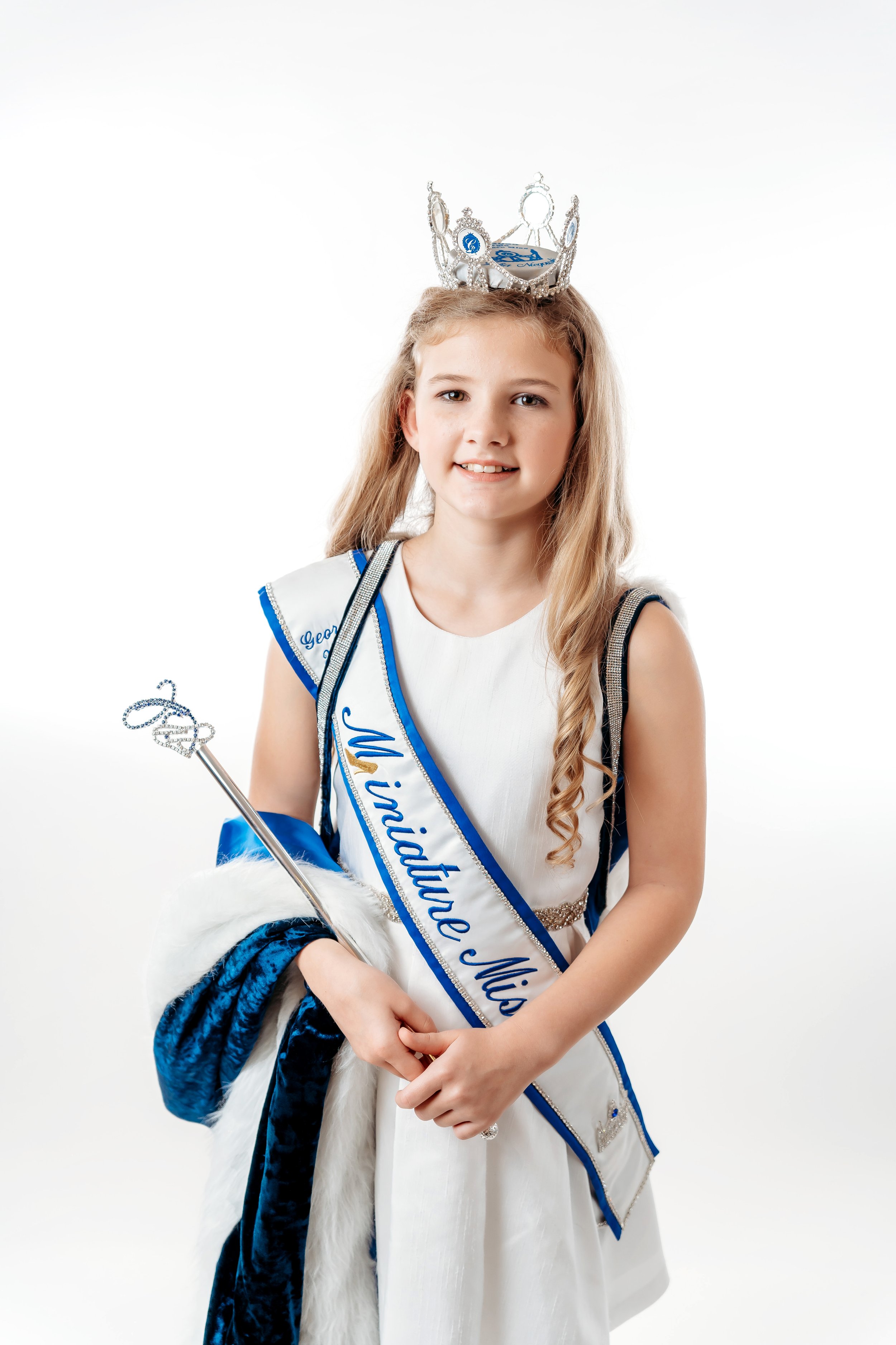 Young girl dressed as a princess with a tiara, wearing a sash that reads 'Missite Miss,' holding a staff and a crown, with a white dress and a royal cape, against a white background.