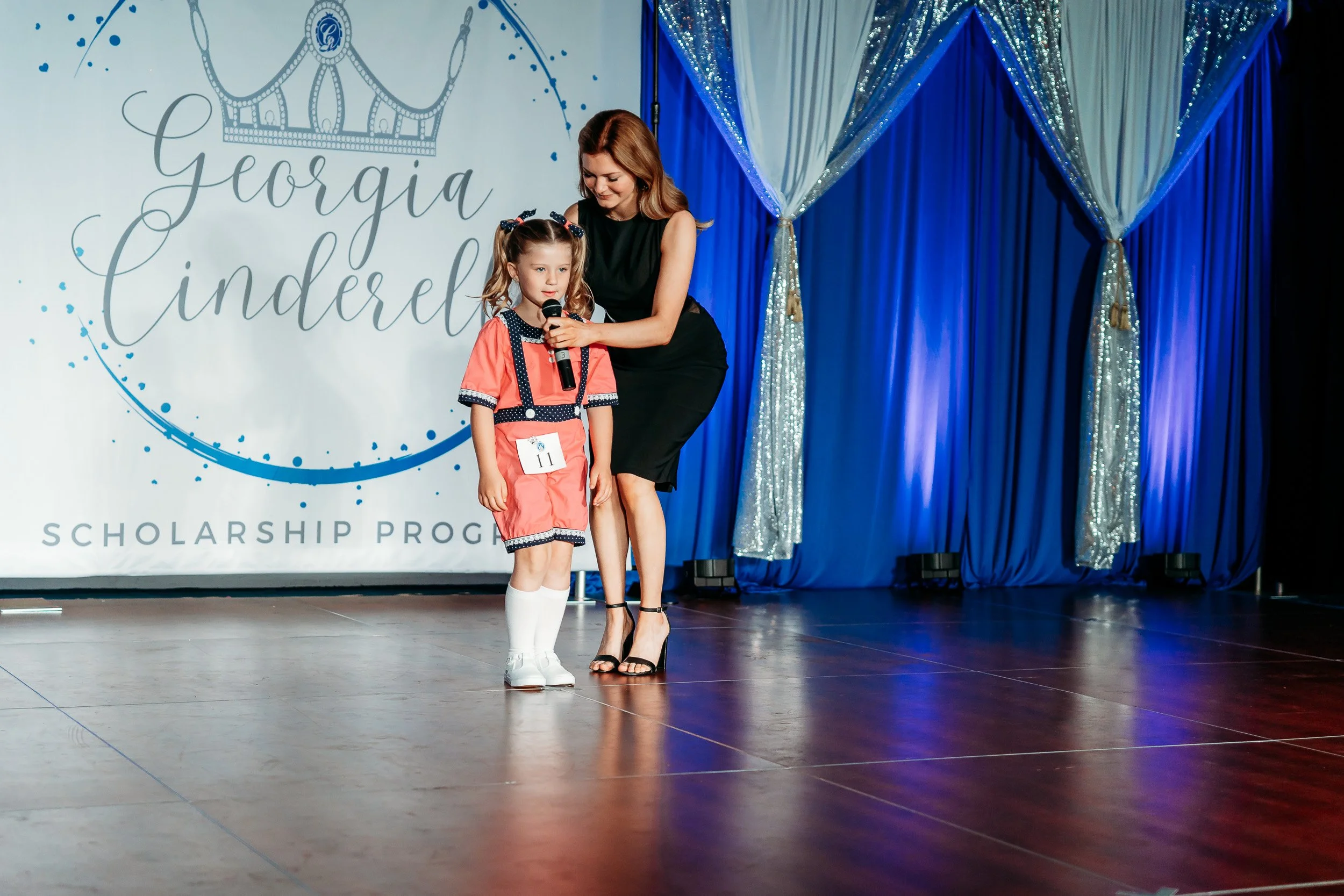Young girl on stage holding a microphone, receiving a speech or award from woman. Background includes a sign that reads 'Georgia Cinderella Scholarship Program' with a crown graphic, blue curtains, and sparkly silver drapes.