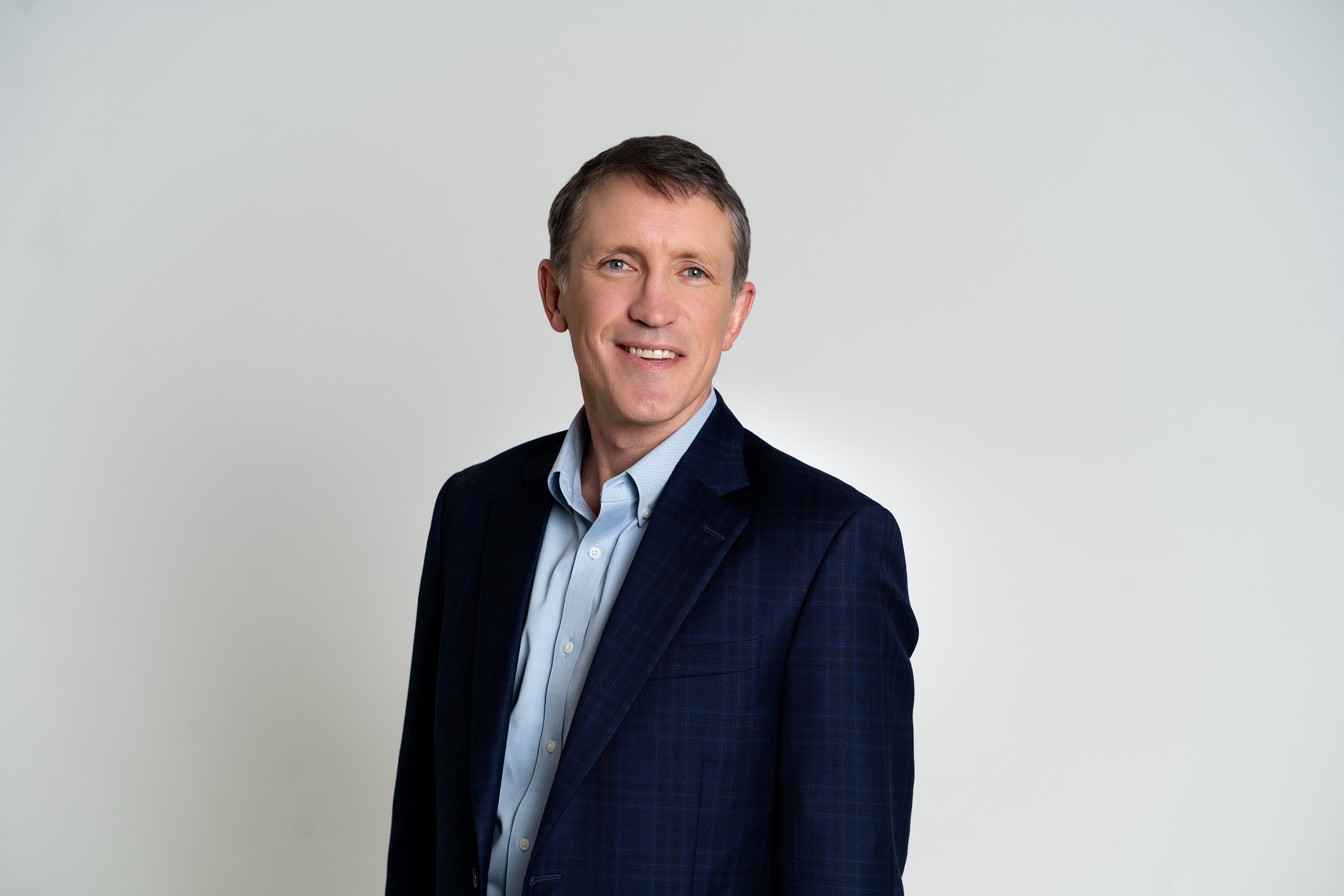 Portrait of a middle-aged man wearing a dark blue blazer and light blue shirt, smiling, against a plain white background.