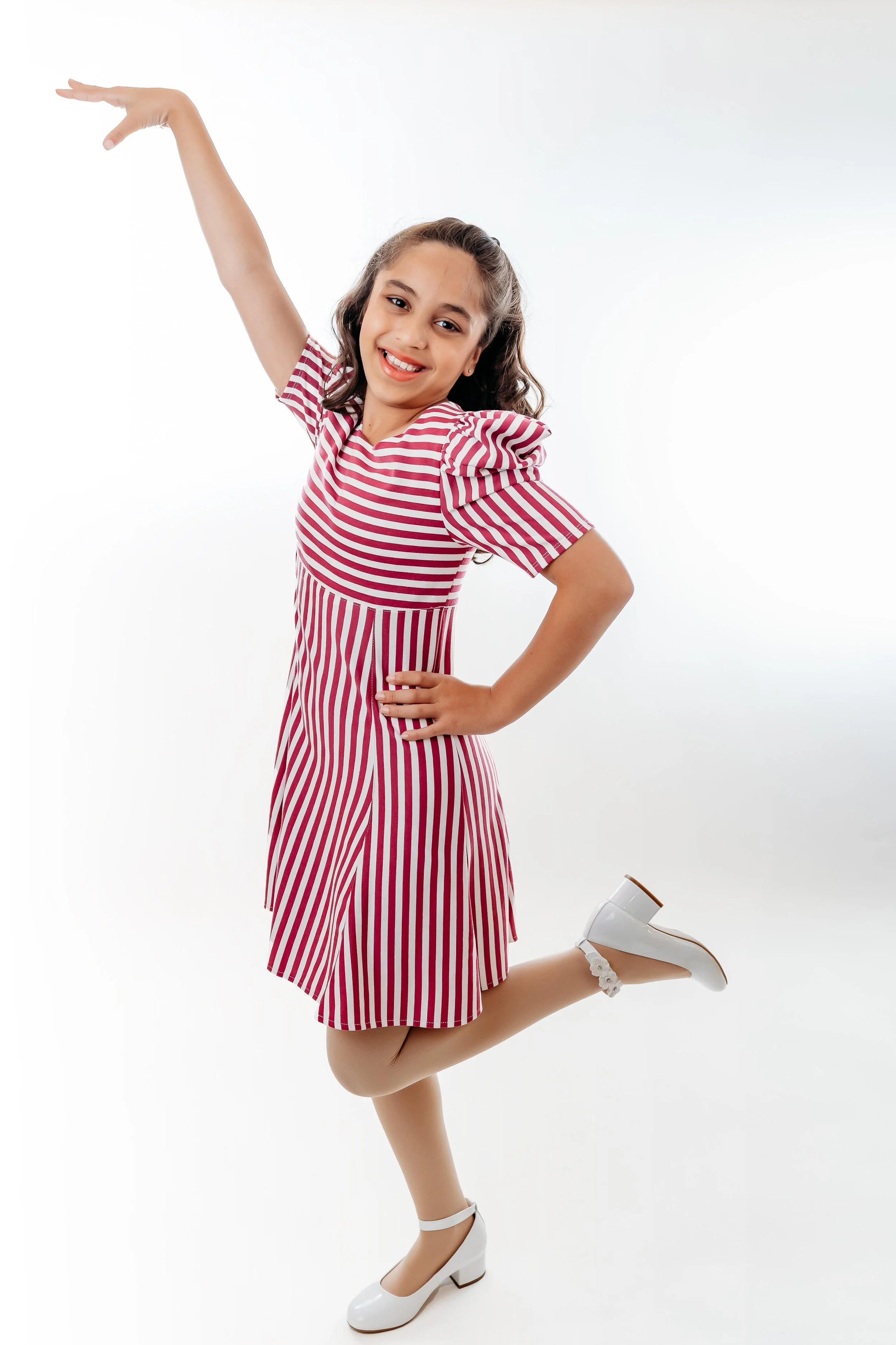 A young girl with long, wavy hair smiling, wearing a red and white striped dress, white shoes, and tan tights, standing on one leg with one arm raised and the other hand on her hip, against a white background.