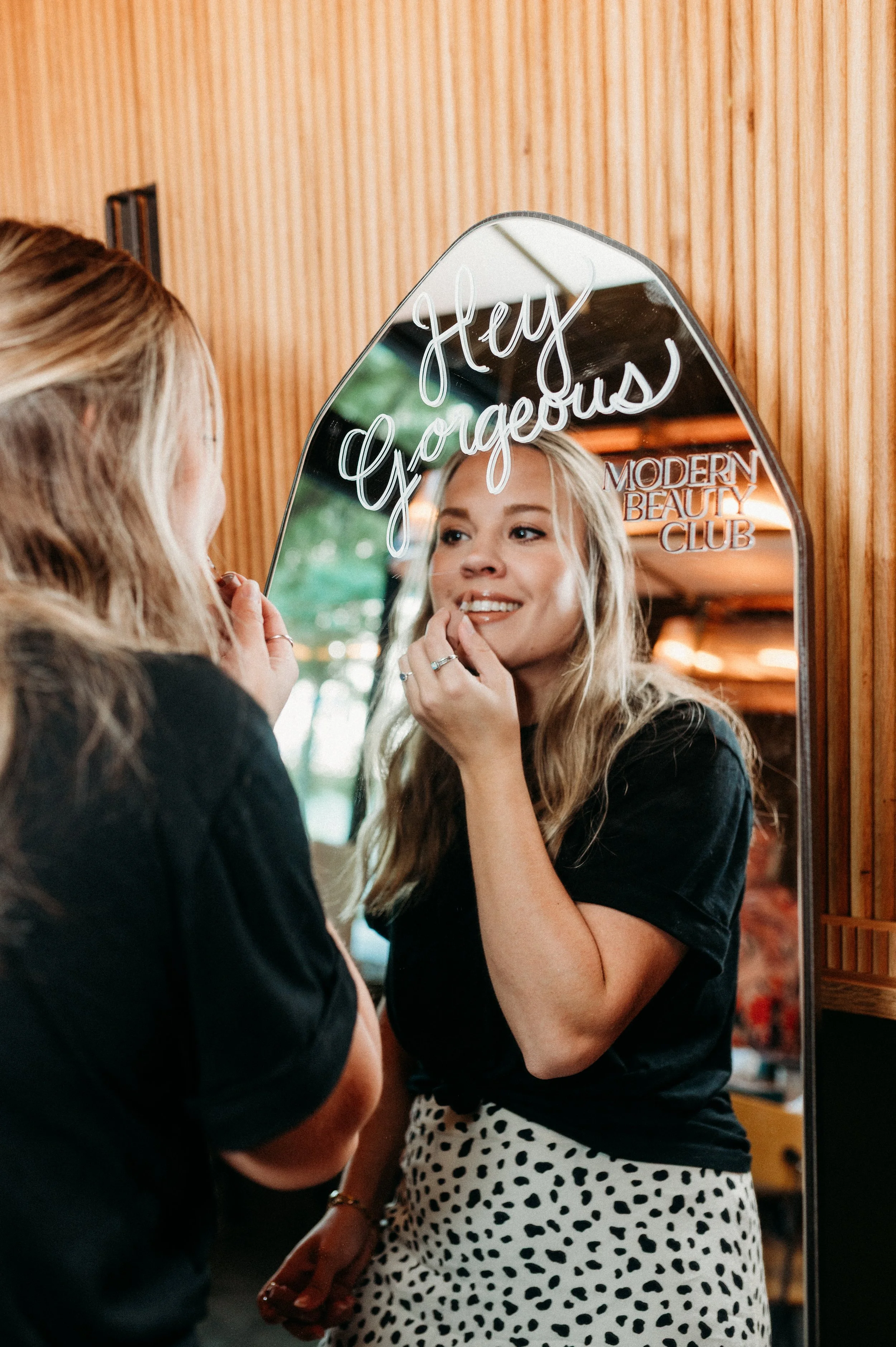 A woman with long blonde hair smiles brightly while looking in a mirror next to writing that says, "Hey Gorgeous."
