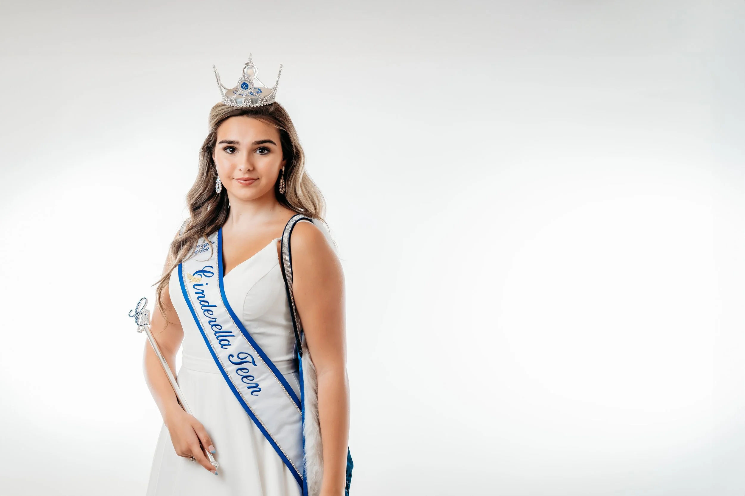 Beauty pageant winner wearing a crown, sash, and white dress, holding a baton, standing against a plain white background.