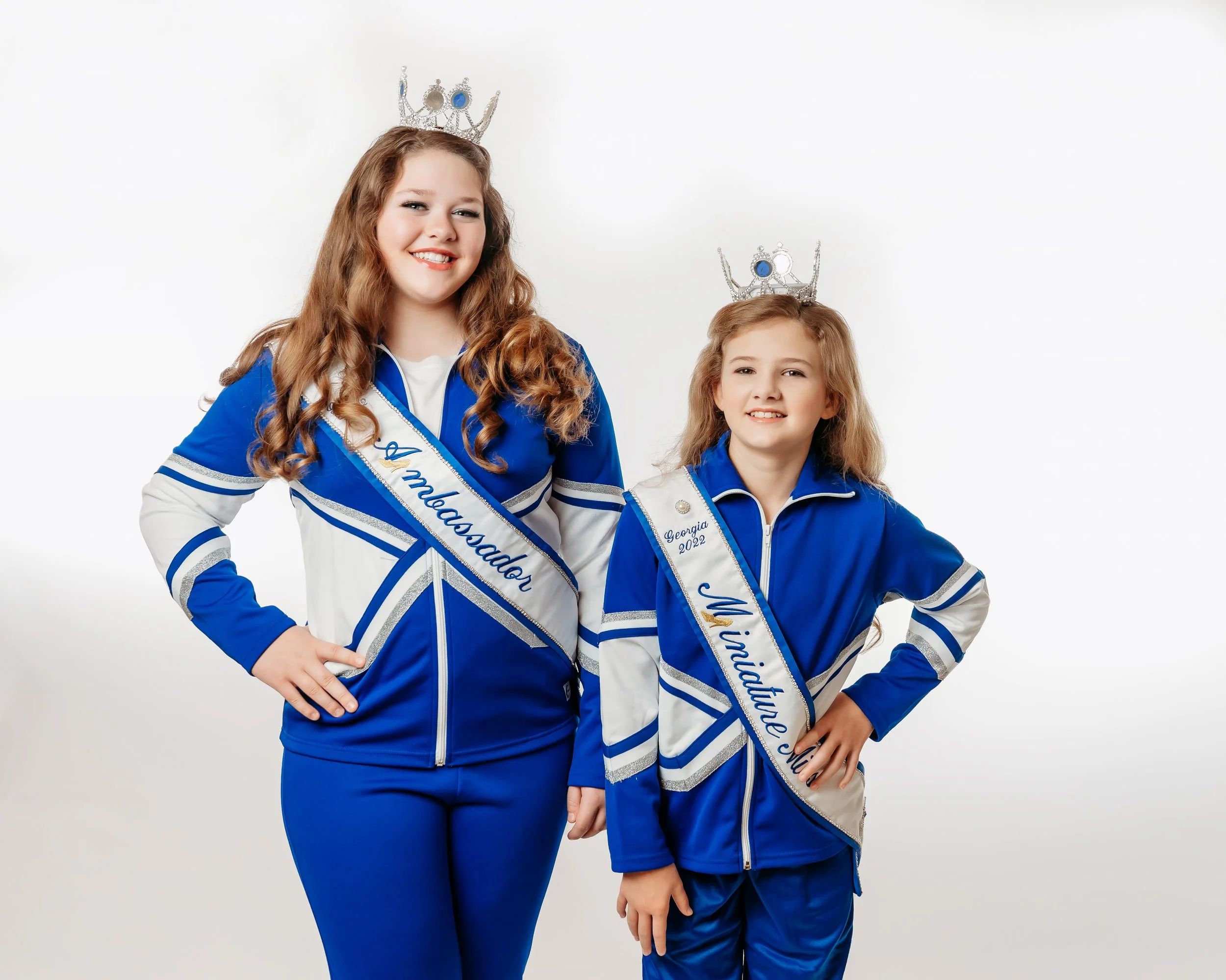 Two young girls wearing blue and white athletic jackets, sashes, and crowns pose against a white background.