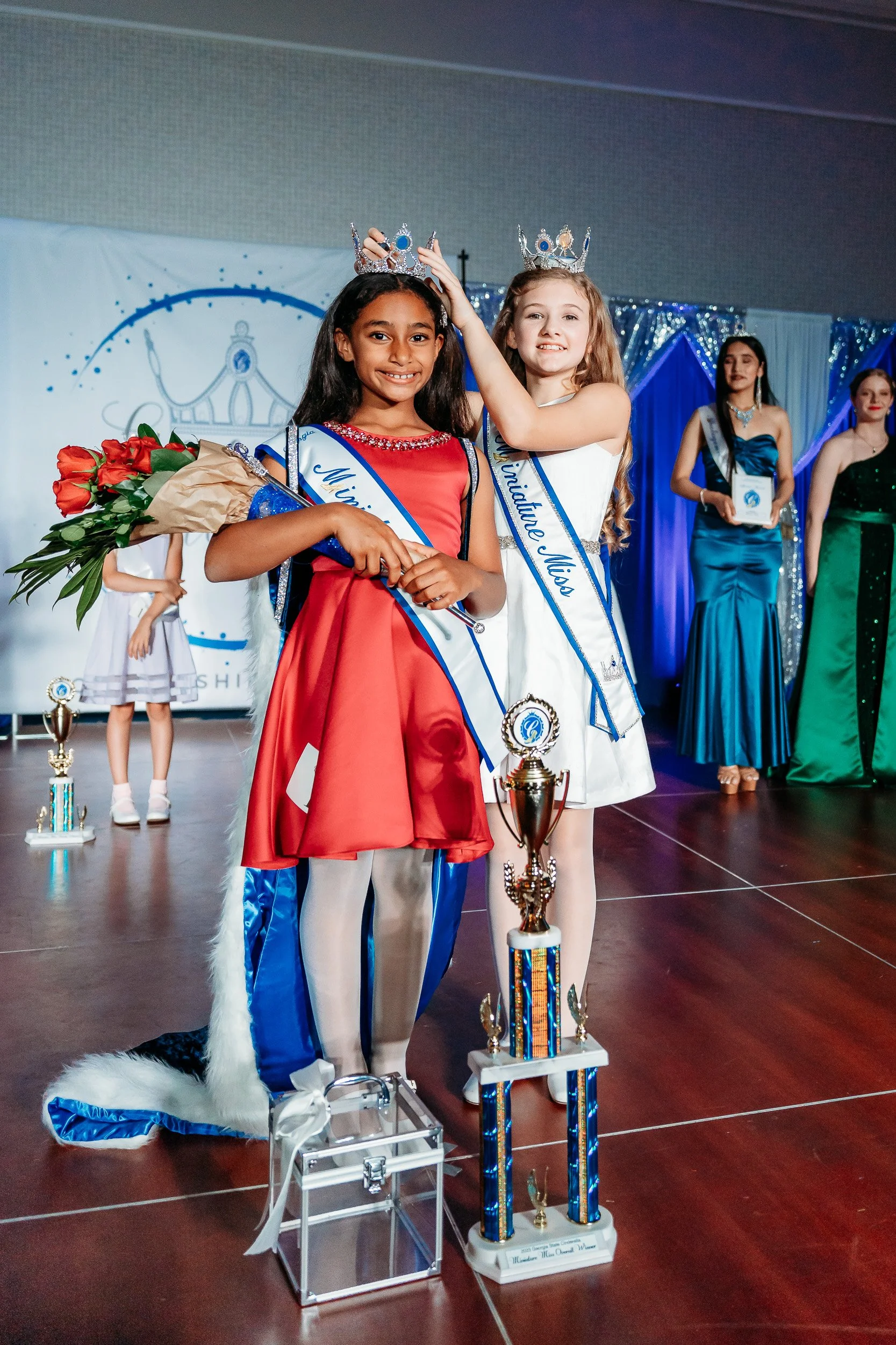 Two young girls wearing sashes and crowns stand on stage, one holding a bouquet of roses and the other adjusting her crown. They are surrounded by other girls in formal dresses, trophies, and awards, at a beauty pageant or similar event.