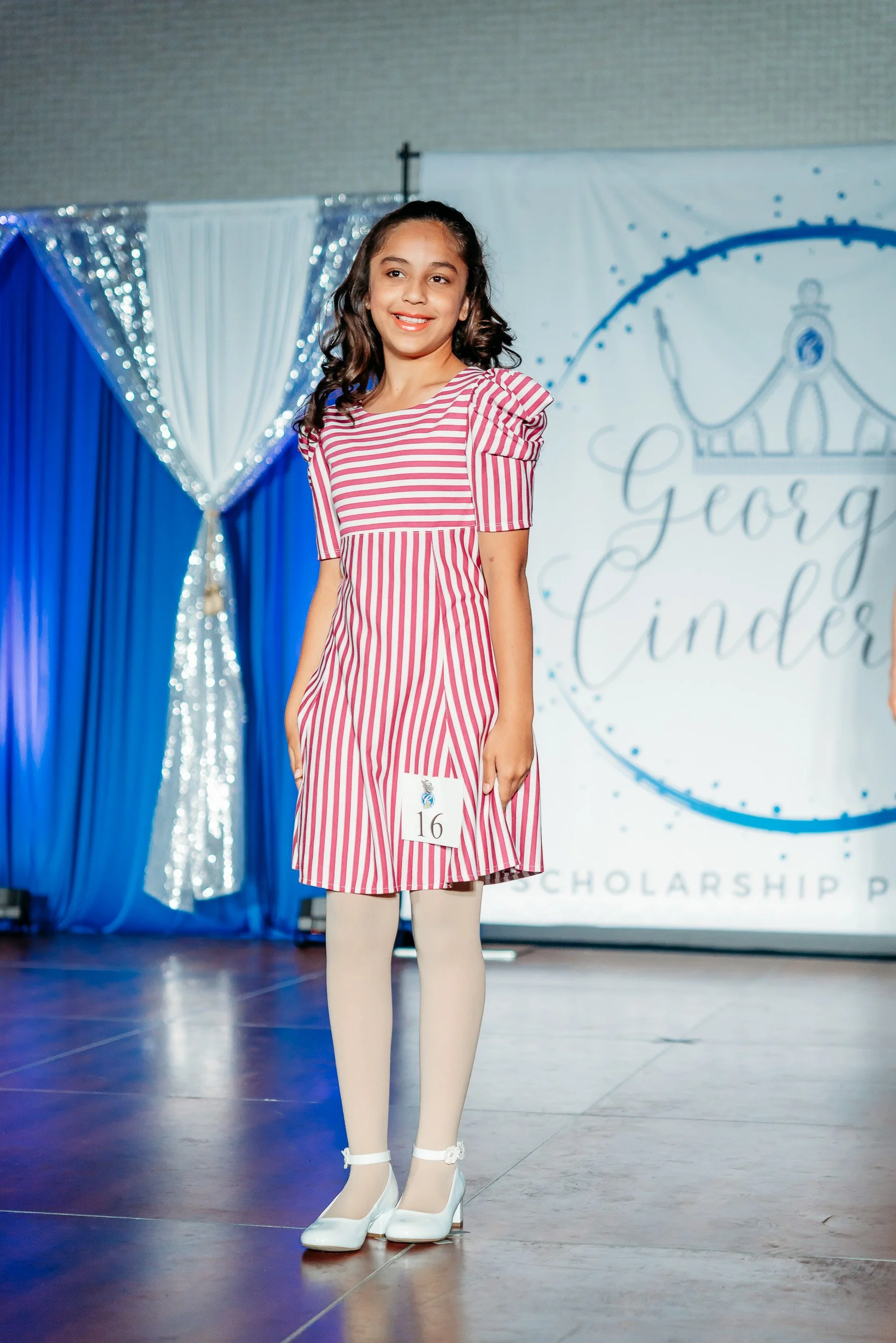 Young girl participating in a pageant wearing a pink and white striped dress with white tights and heels, standing on stage in front of a backdrop with the words 'George Cinder' and a crown illustration.