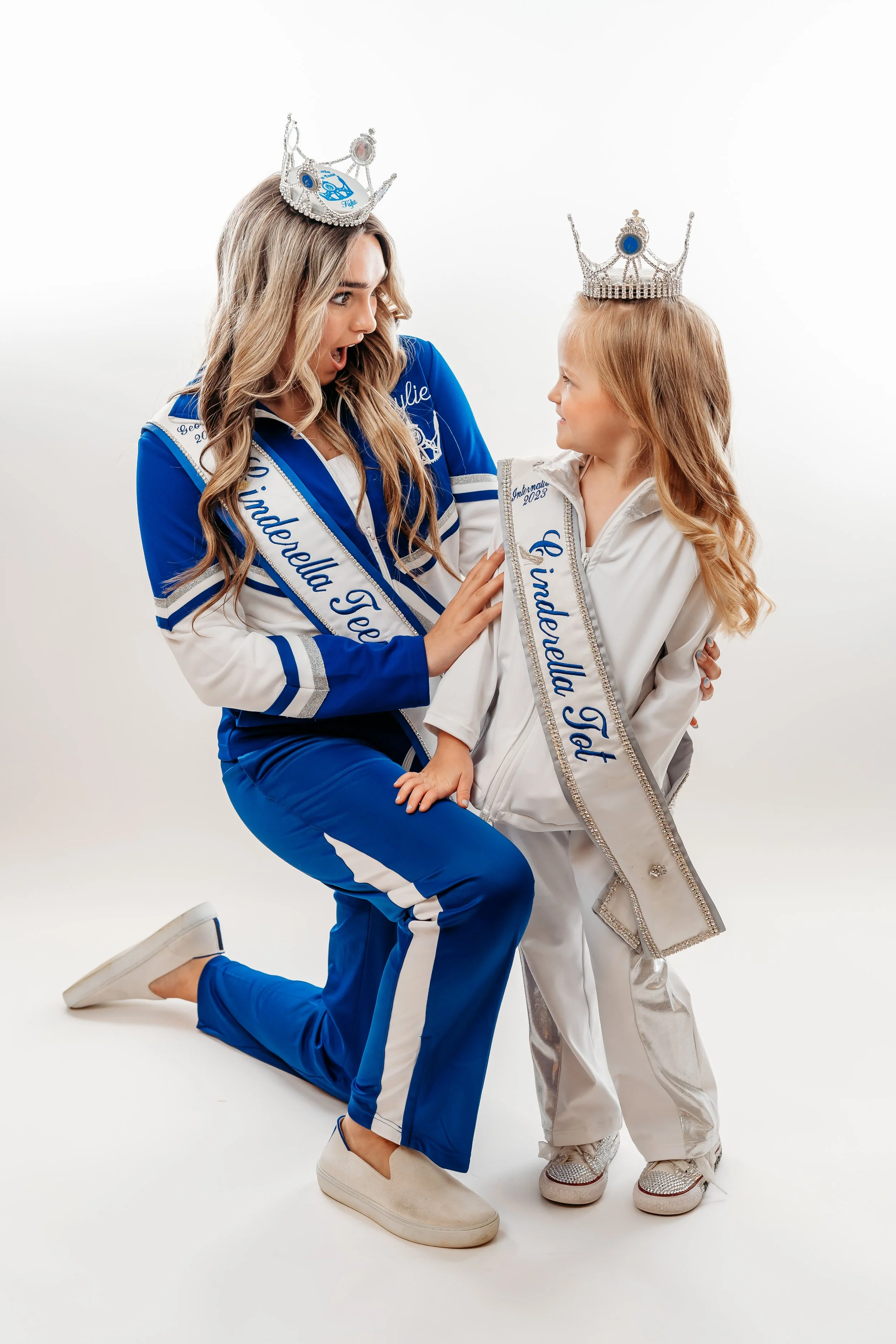 A woman and a young girl wearing crowns and sashes that say "Miss Geodebella 2023" and "Geodebella Jr. 2023". The woman is kneeling, looking surprised, while the girl stands, smiling. The woman has long wavy hair, is dressed in a blue and white track