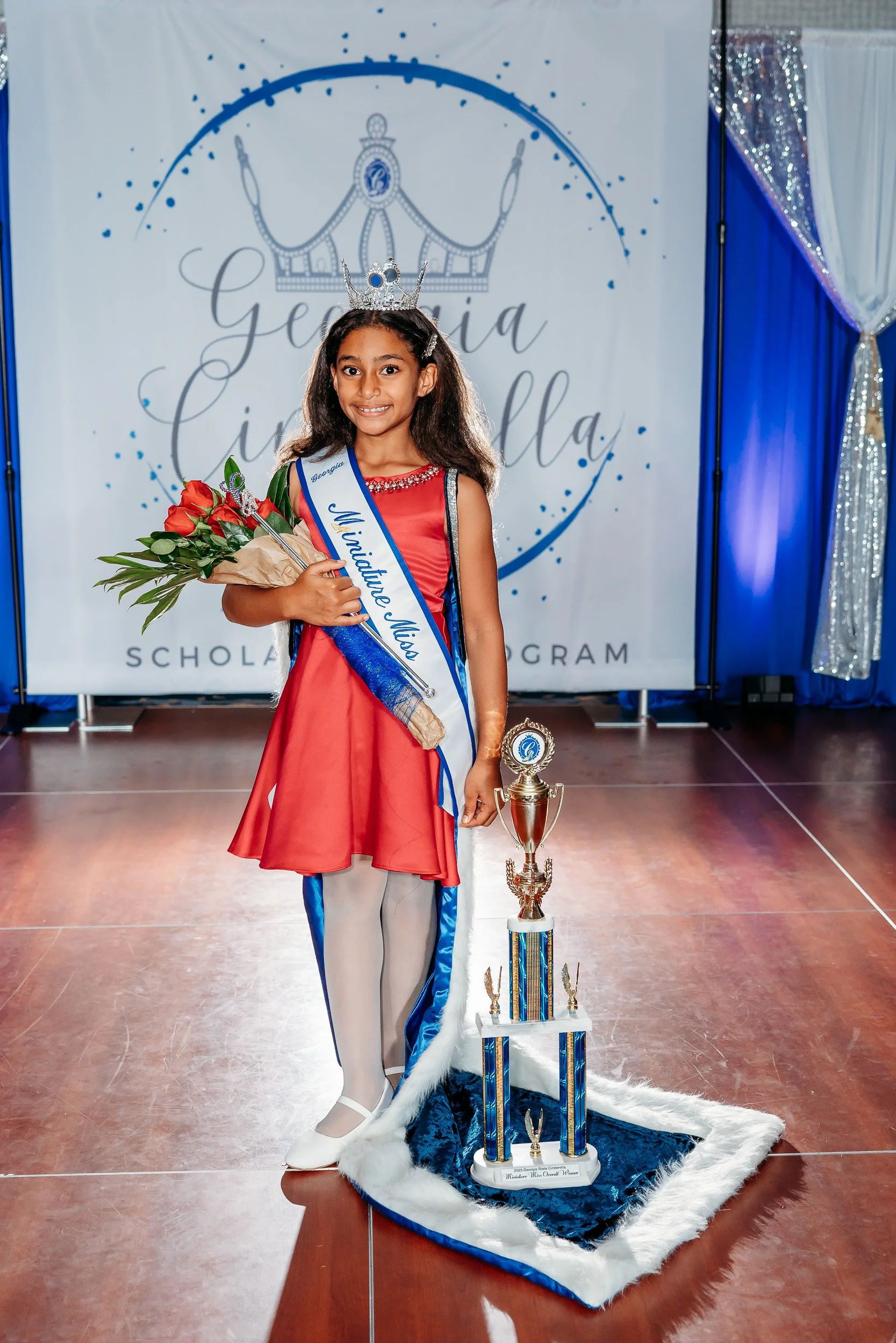 A young girl wearing a red dress, a silver tiara, and a sash reading "Miss Georgia" stands on a stage holding a bouquet of red roses and a trophy. Behind her is a festive backdrop with a crown graphic and the words "Georgia Cinderella". The floor is 
