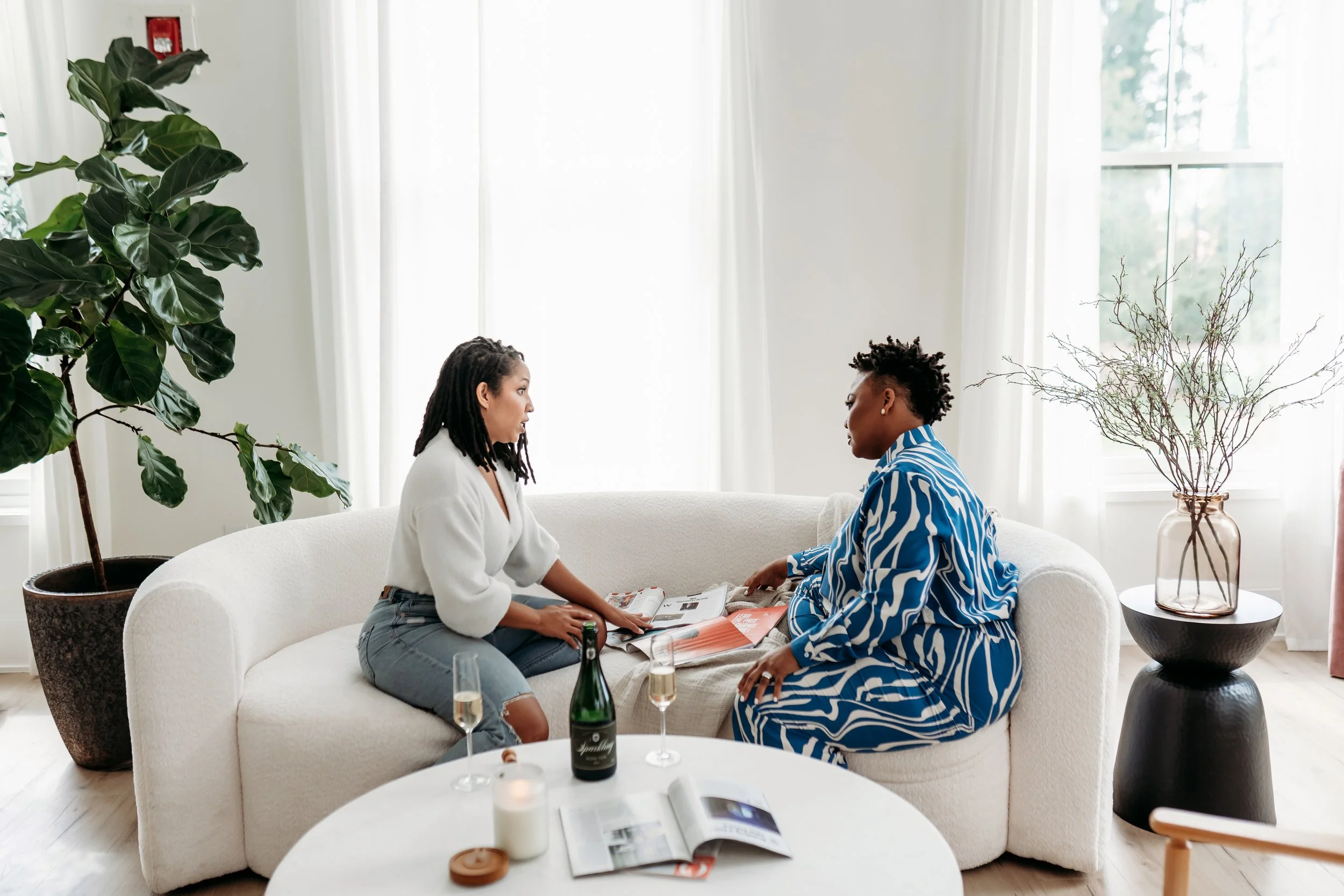 two women sitting on white couch in white studio looking at magazines for brand photo session