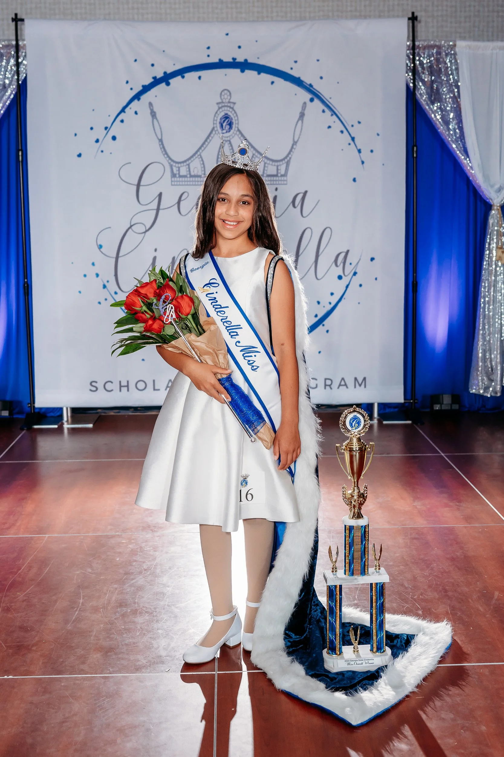 Young girl wearing a Crown and sash that reads "Gwedealla Miss", holding a bouquet of red roses, standing next to a large trophy on the floor, at a pageant event with a decorated backdrop that says "Georgia" and "Little Miss".