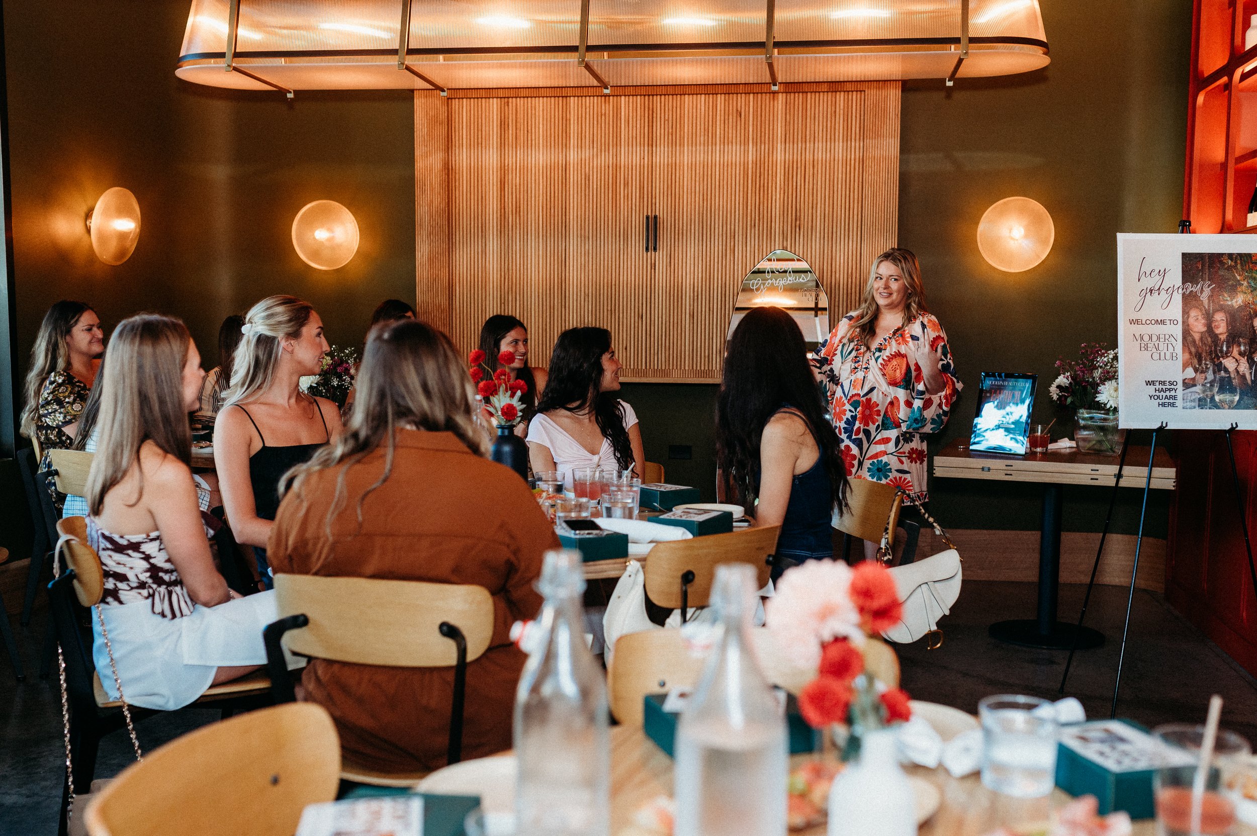 A woman in a patterned dress stands speaking to a group of seated people in a warmly lit room.