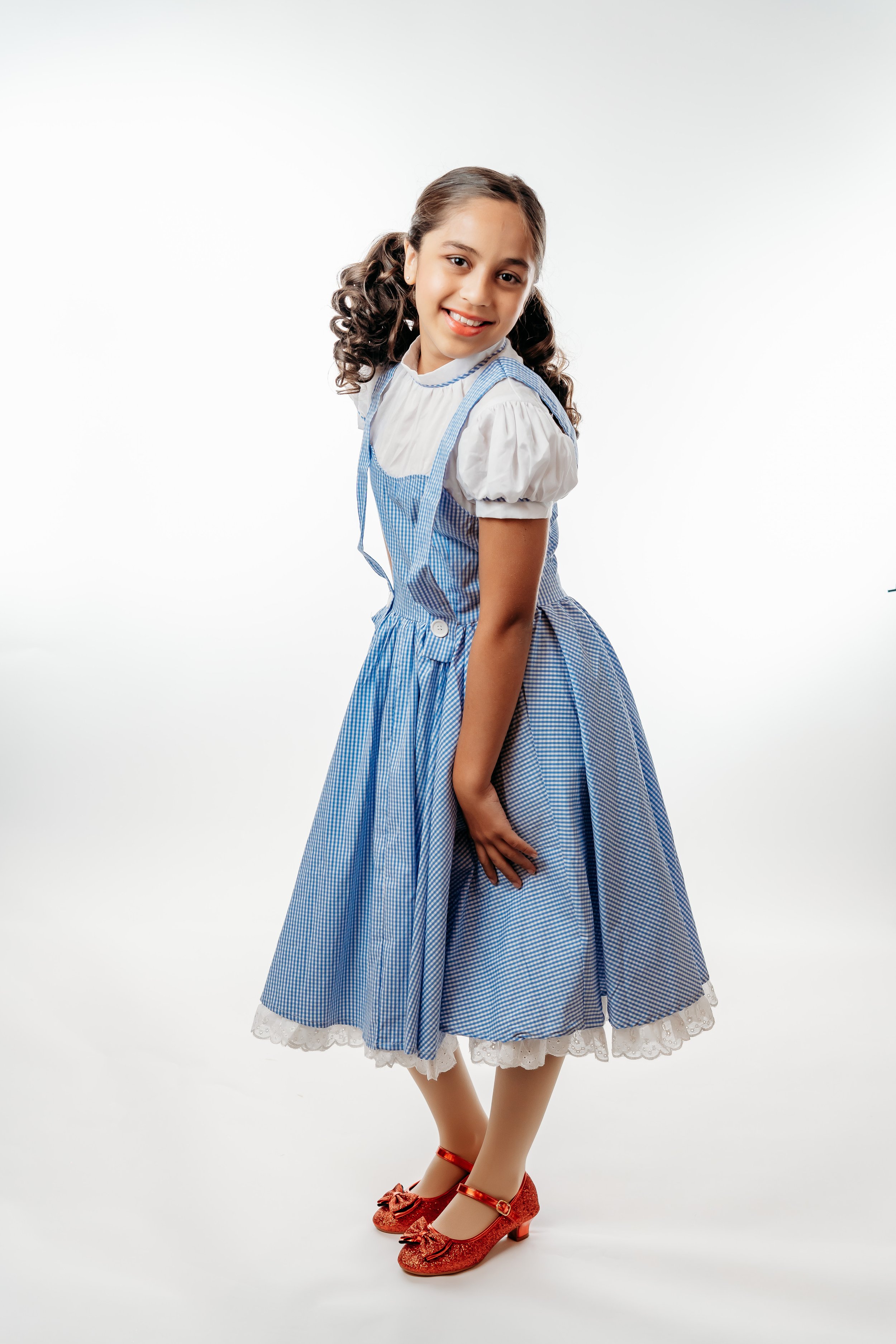 Young girl with curly hair smiling, wearing a blue and white gingham dress with puffy sleeves, a white blouse underneath, and red glittery shoes with bows, standing against a white background.