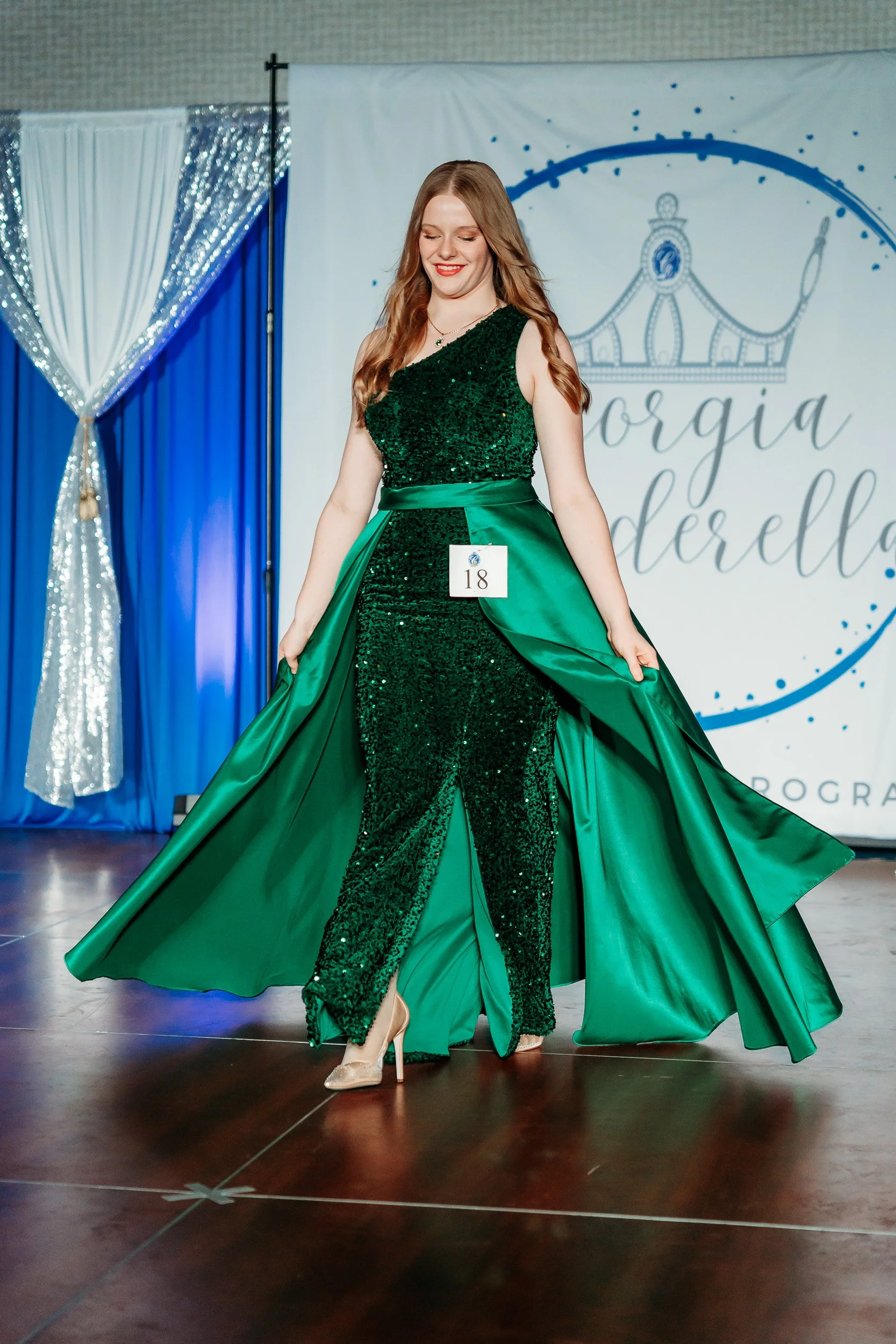 A woman in a glittery emerald green gown with a matching satin overskirt standing on a stage during a pageant or fashion show, with a backdrop showing a crown and script writing.