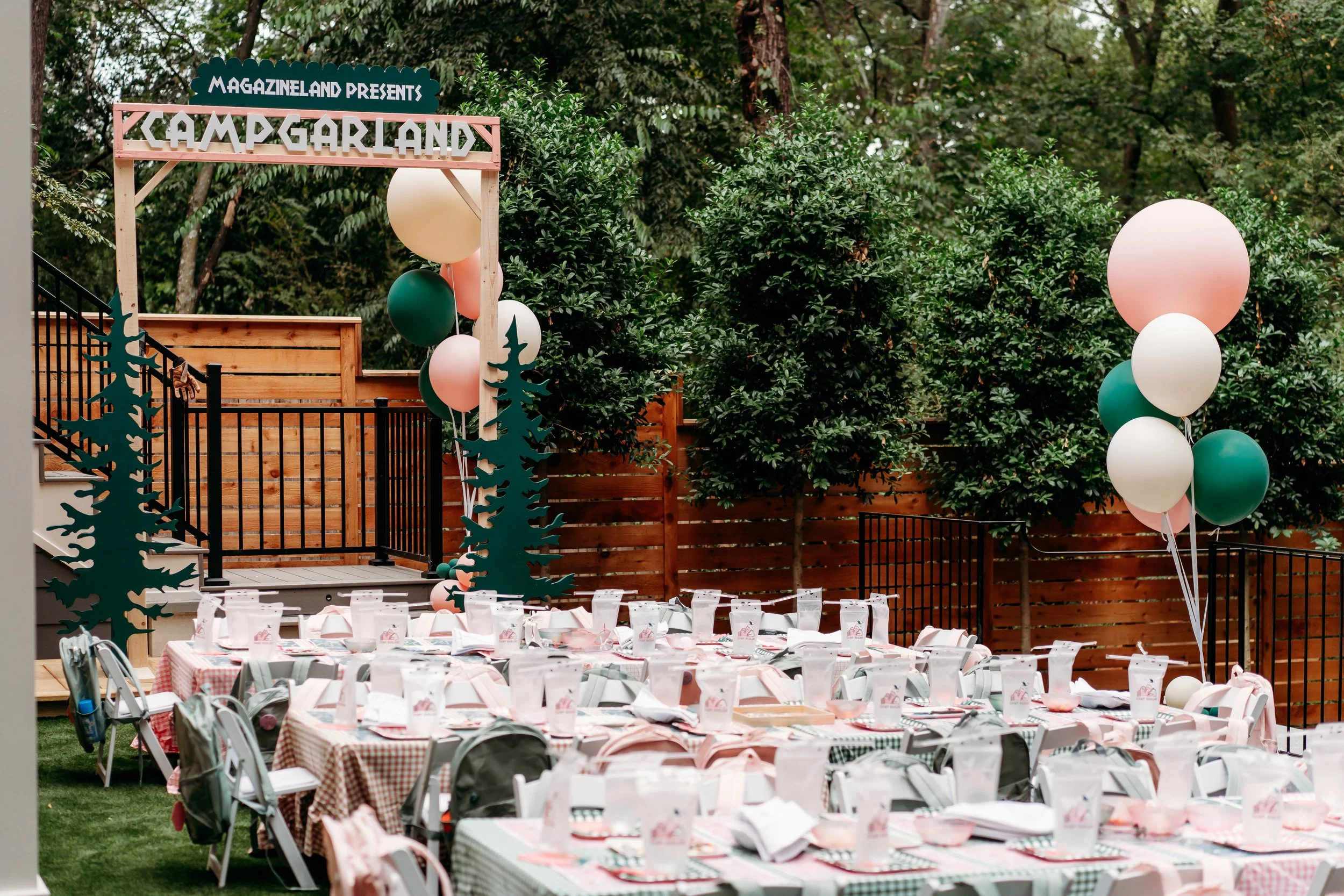Outdoor party setup with white tables and chairs, surrounded by green trees. Balloons in peach, white, and green add a festive touch.