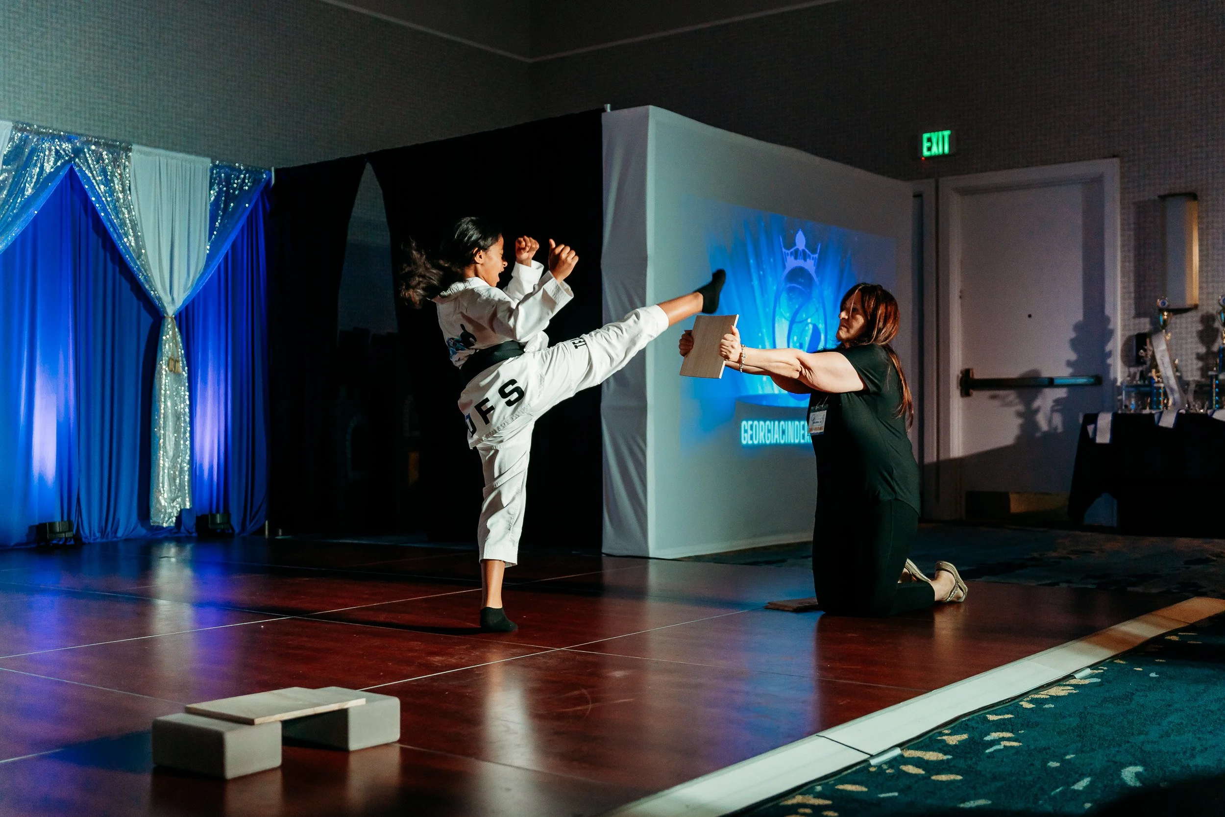 A girl in a white martial arts uniform performing a high kick towards a woman kneeling on the stage with a kickpad during a martial arts demonstration at an event.