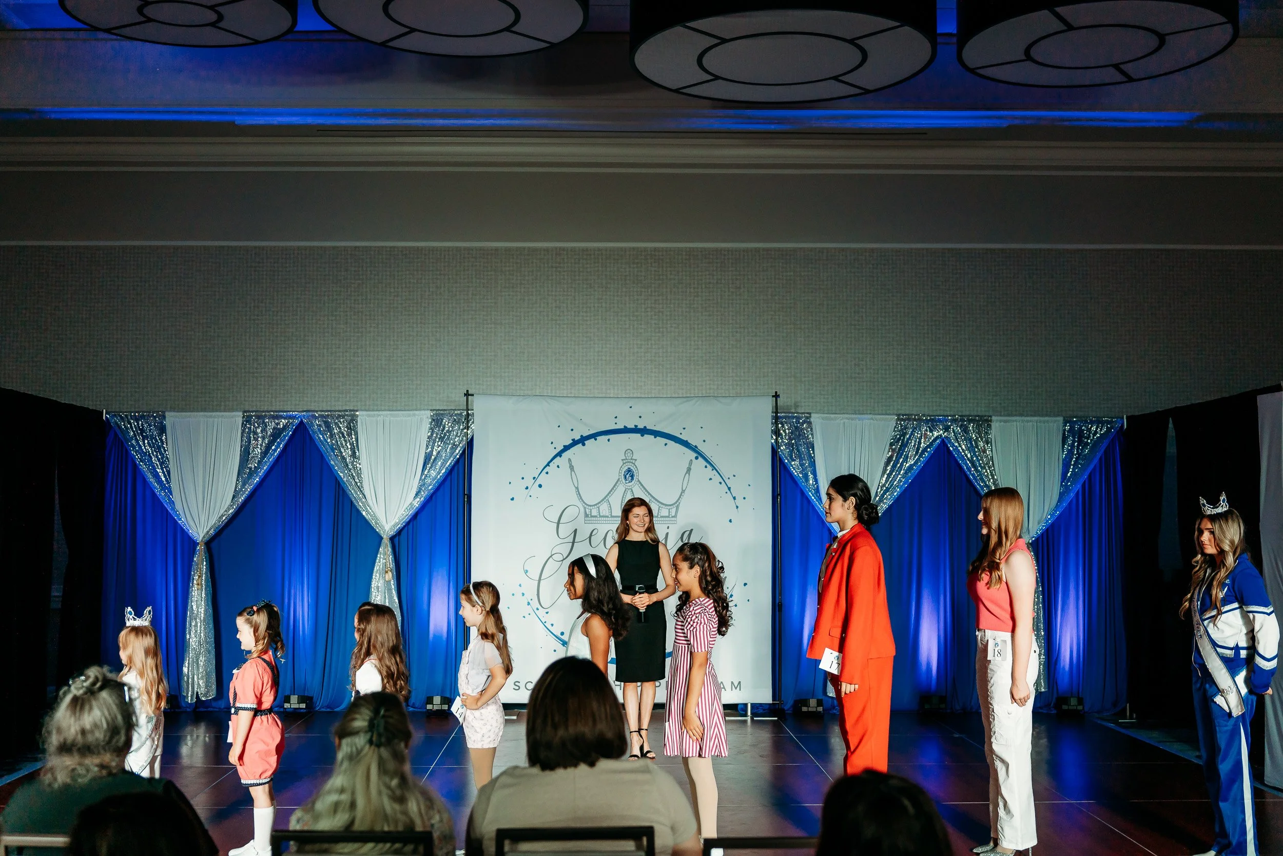 Children participating in a beauty pageant on stage, with three women judges or organizers, and a backdrop with elegant blue and white drapery and a logo.
