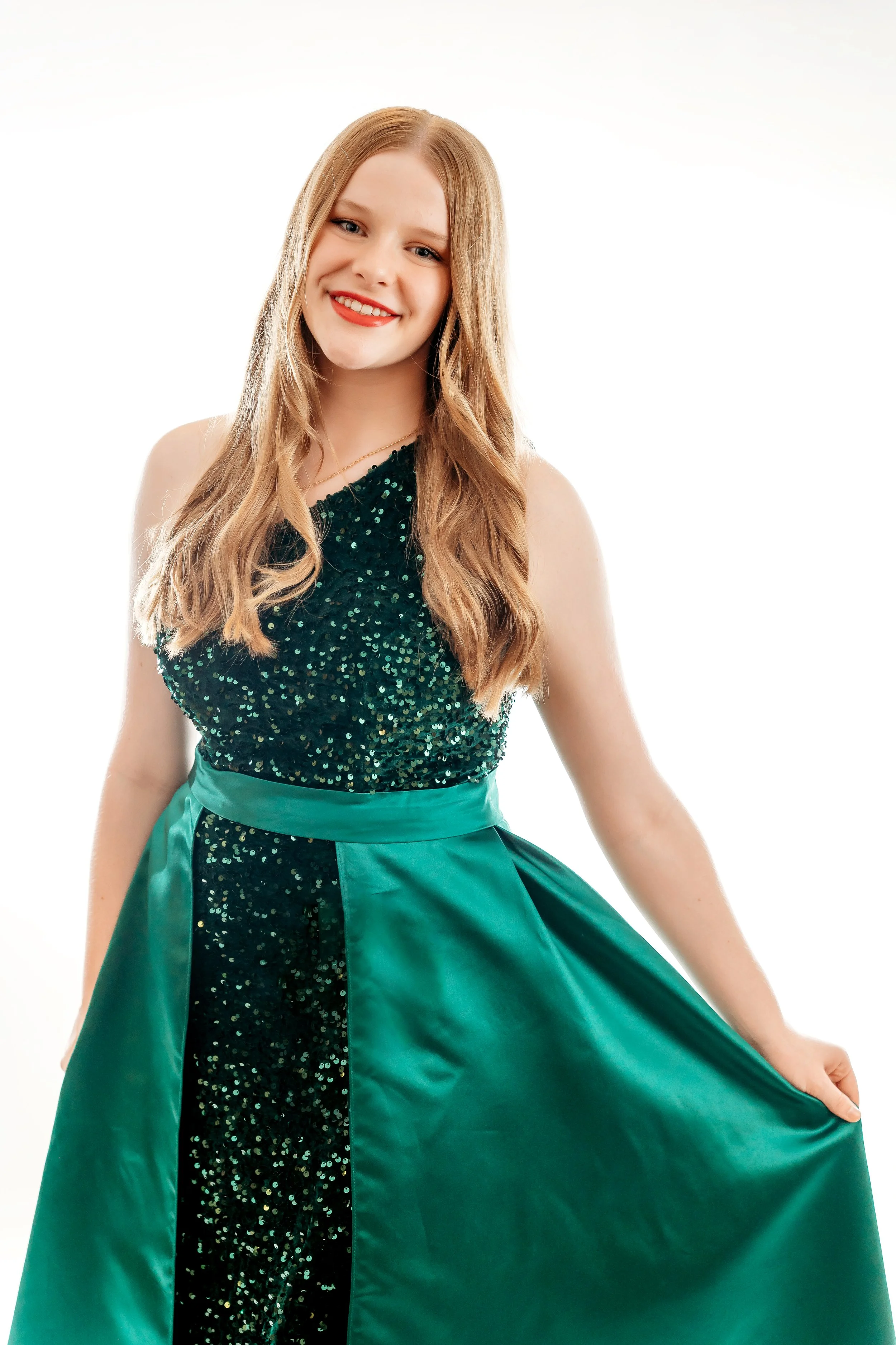 A young woman with long, wavy blonde hair wearing a dark green, sequined sleeveless top and a matching satin skirt with a dark green sequined stripe down the center, posing in front of a plain white background.