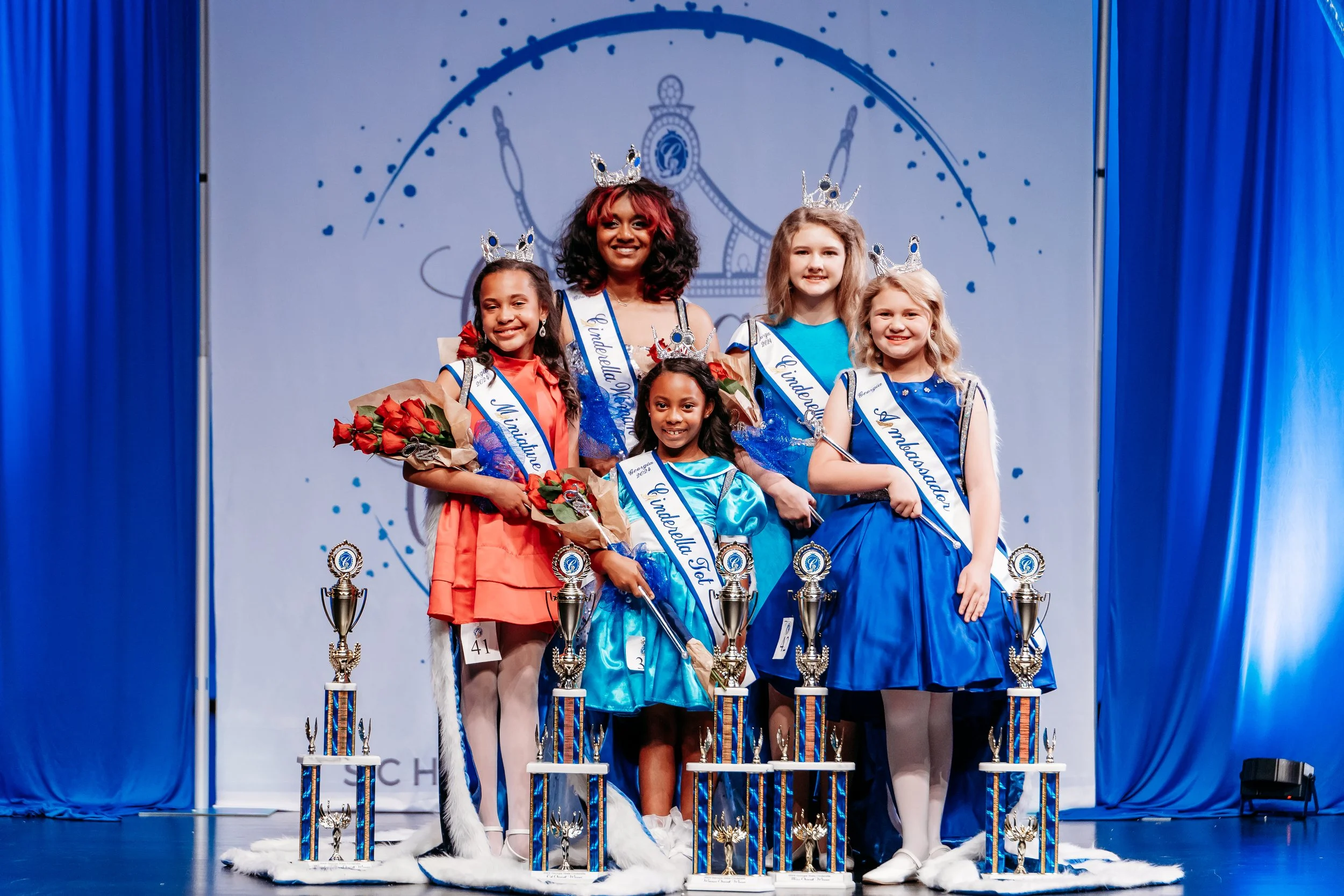 A group of five young girls and one woman on stage at a pageant, all wearing sashes and crowns, holding flowers, with trophies in front of them. The background features a large logo and blue curtains.